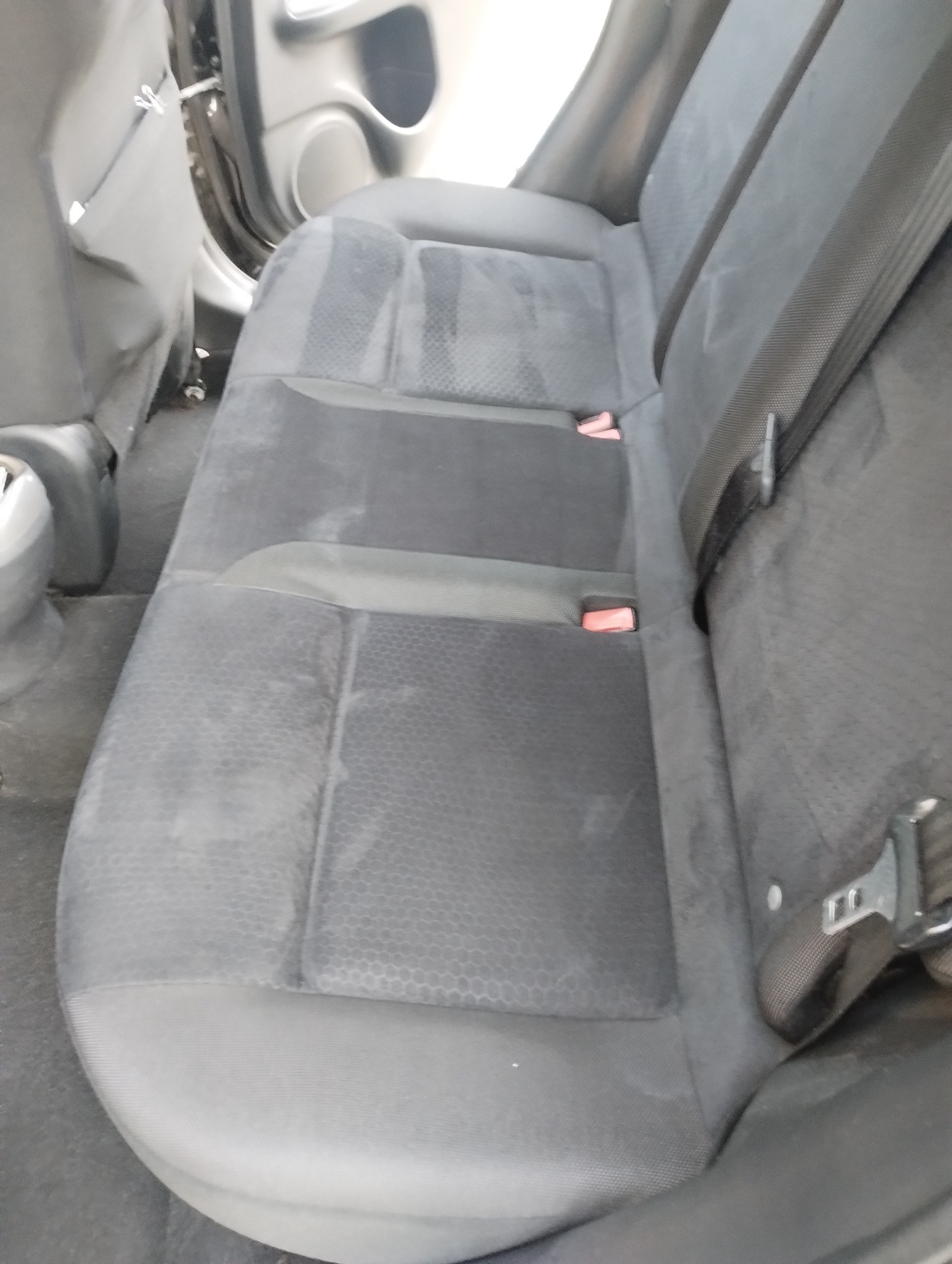 Szara, materiałowa tylna kanapa samochodowa z widocznymi plamami i pasami bezpieczeństwa, wymagająca odświeżenia. Widok z góry, wnętrze pojazdu.