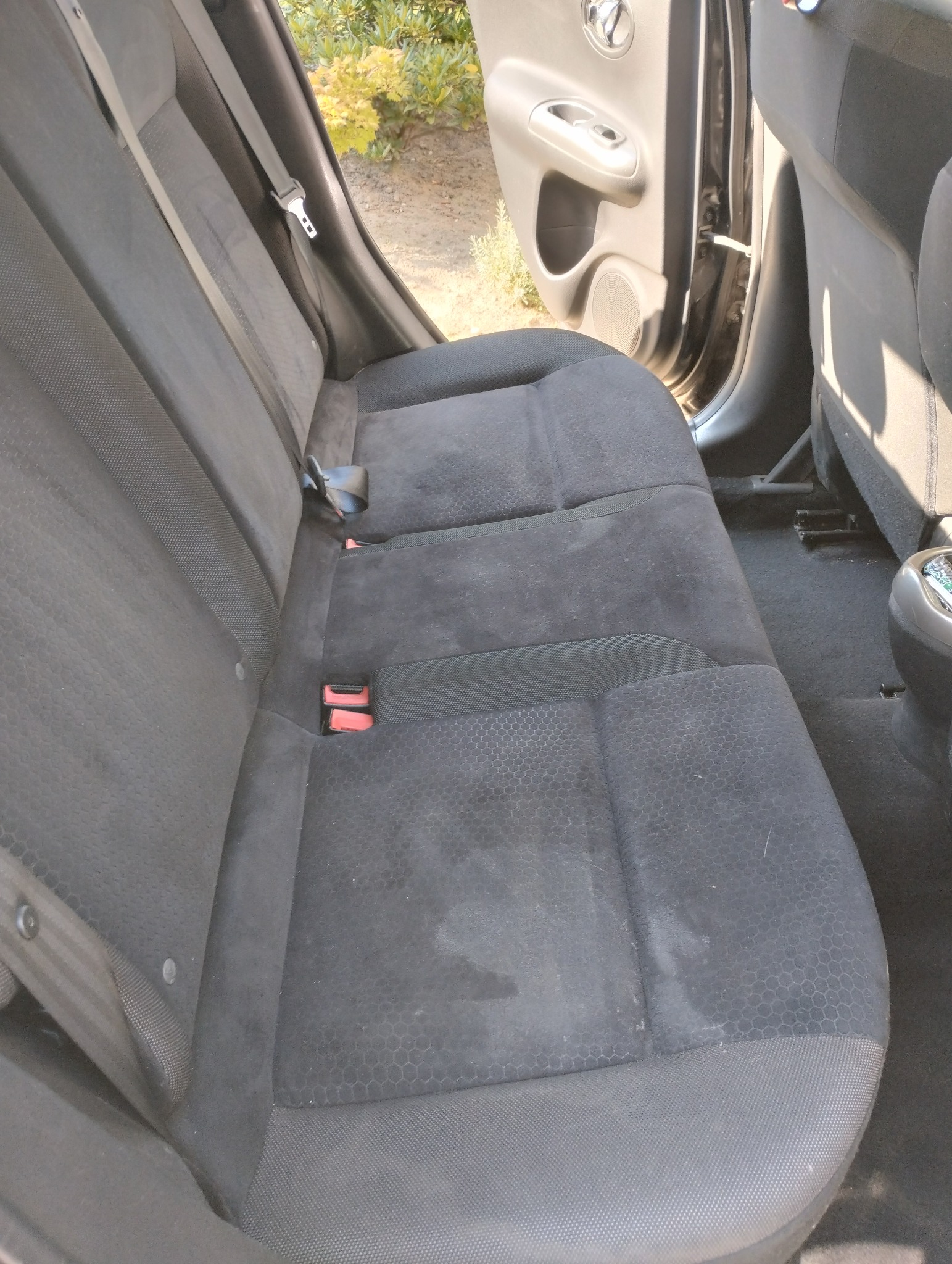 Ciemna, materiałowa tylna kanapa samochodowa z widocznymi plamami i zabrudzeniami, pasami bezpieczeństwa. Widoczne elementy drzwi i podłogi auta.