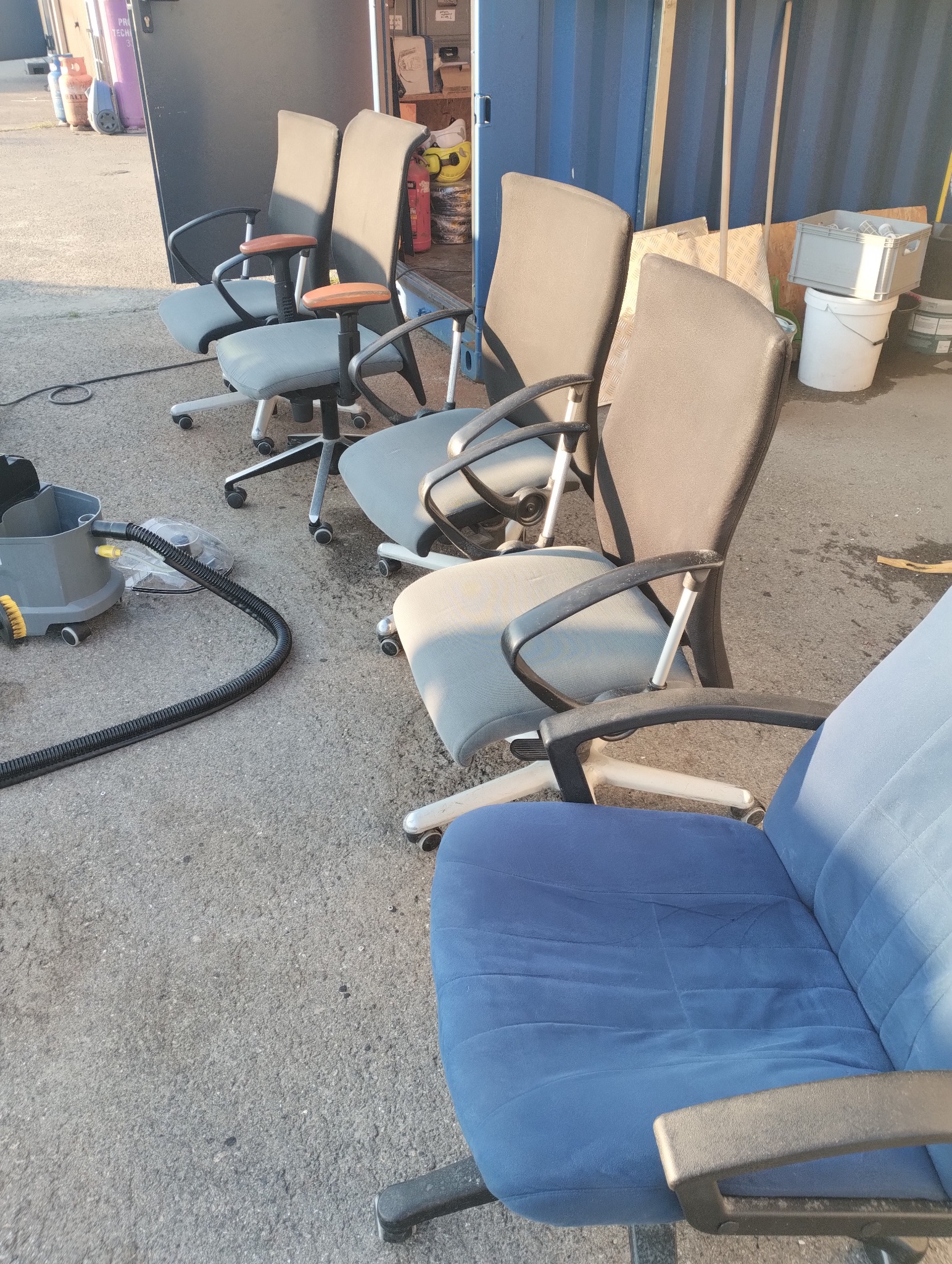 Rząd krzeseł biurowych na kółkach, częściowo tapicerowanych, ustawionych na zewnątrz obok sprzętu czyszczącego, sugerujący przygotowanie do czyszczenia tapicerki.
