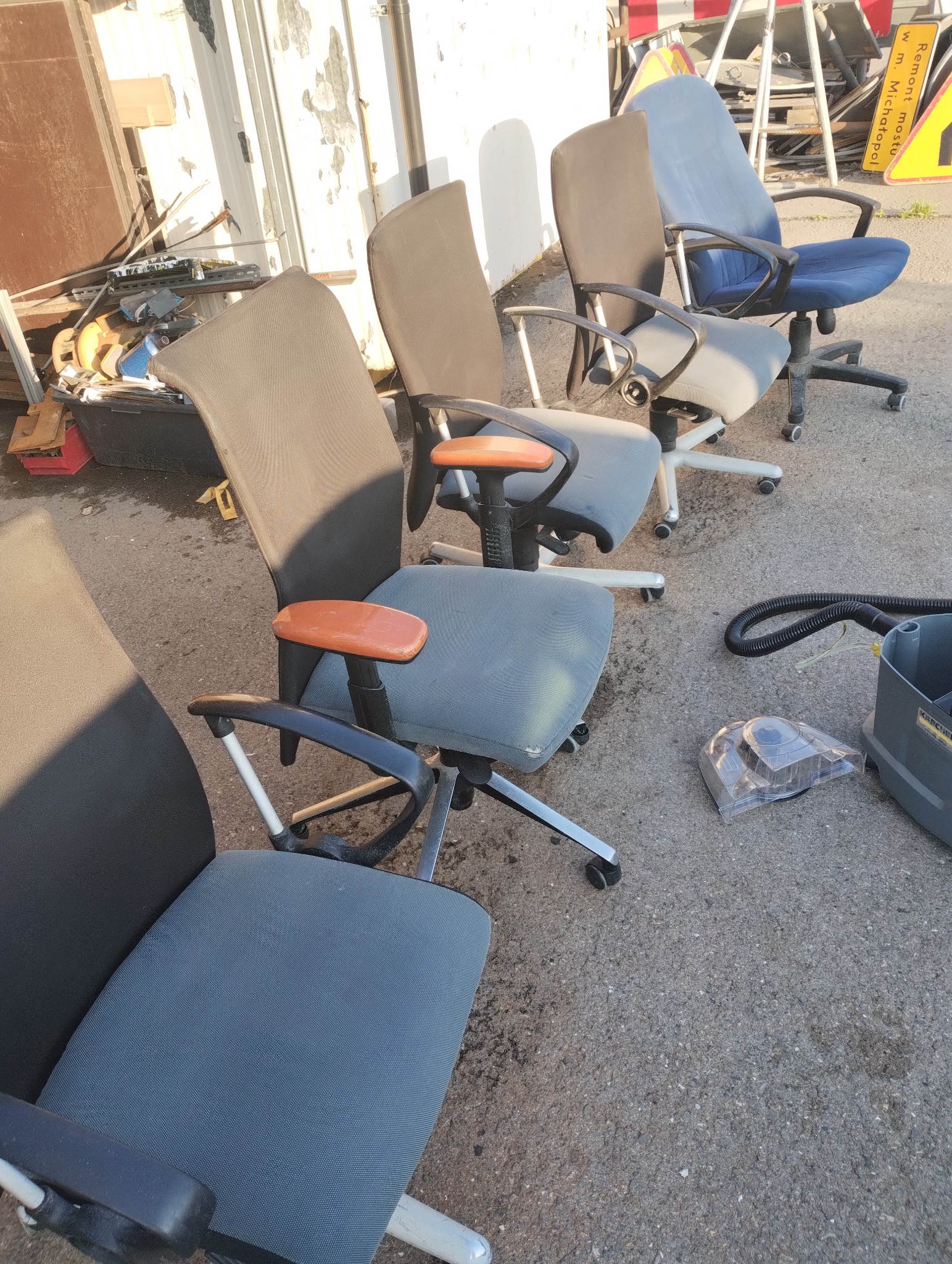 Rząd używanych, biurowych krzeseł na kółkach, obok sprzęt do prania tapicerki. Widoczne ślady użytkowania. Przygotowanie do czyszczenia.