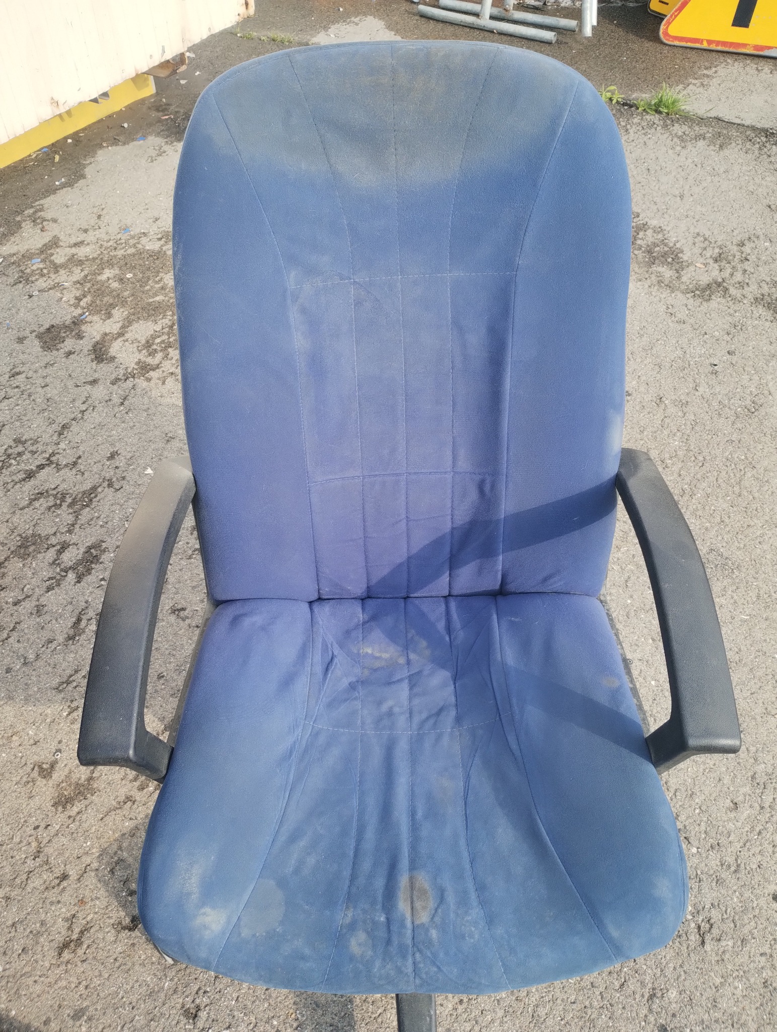 Niebieskie krzesło biurowe z plamami na tapicerce, widoczne ślady użytkowania. Ujęcie z przodu, na zewnątrz, na tle betonowej nawierzchni i fragmentu metalowego ogrodzenia.