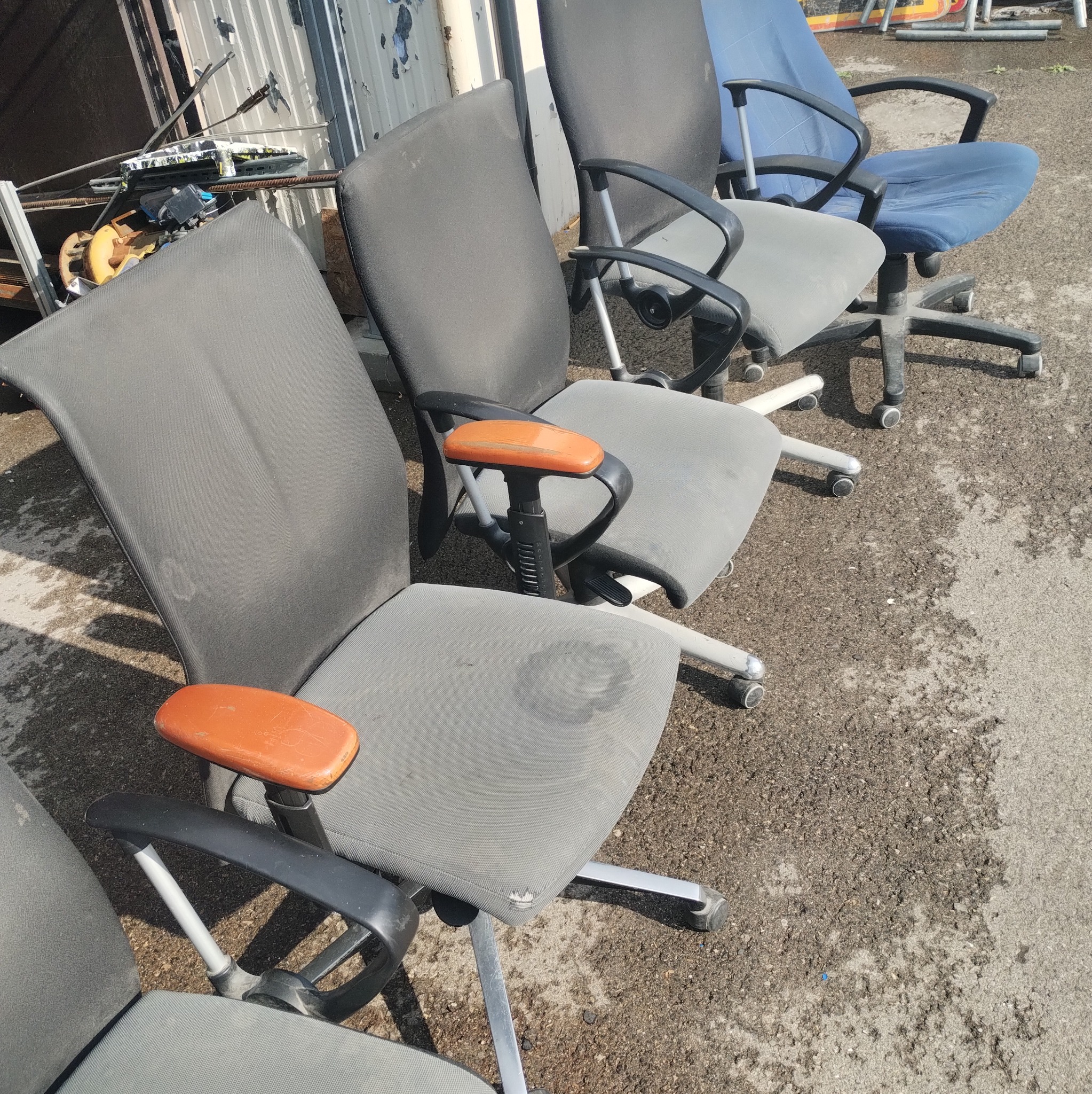Rząd używanych, biurowych krzeseł obrotowych z widocznymi plamami na tapicerce, ustawionych na zewnątrz. Krzesła w odcieniach szarości i błękitu, z elementami pomarańczowymi.