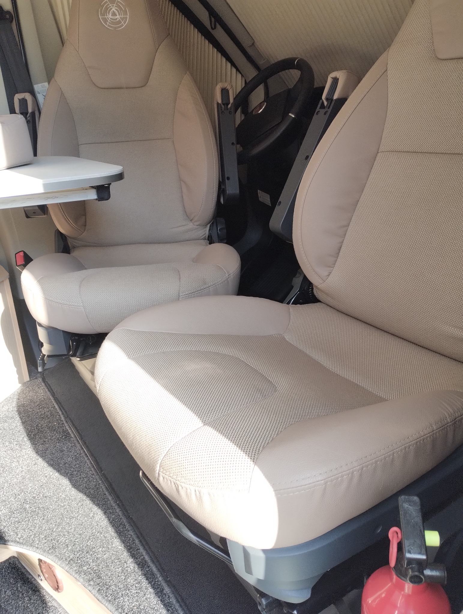 Beżowe, tekstylne fotele kierowcy i pasażera w kamperze, widok z góry. Na podłodze szara wykładzina. Widoczna kierownica i stolik.