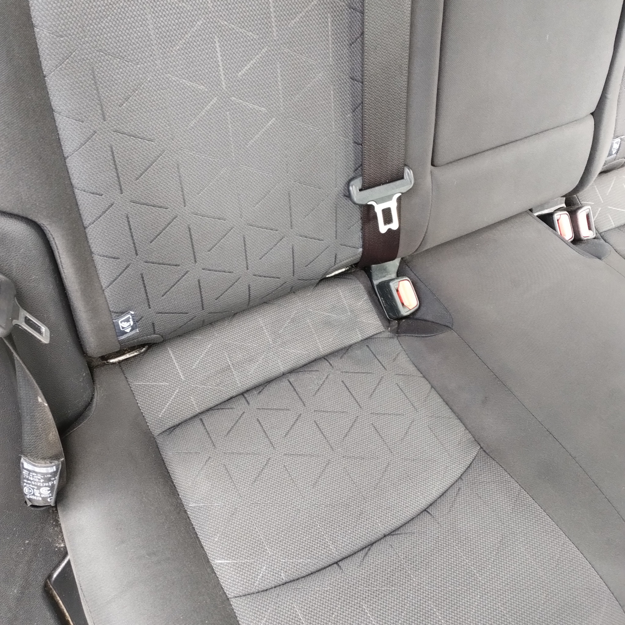 Szare, tekstylne siedzenie samochodowe z pasami bezpieczeństwa. Widoczna faktura materiału i detale mocowań pasów. Czyste, zadbane wnętrze pojazdu.