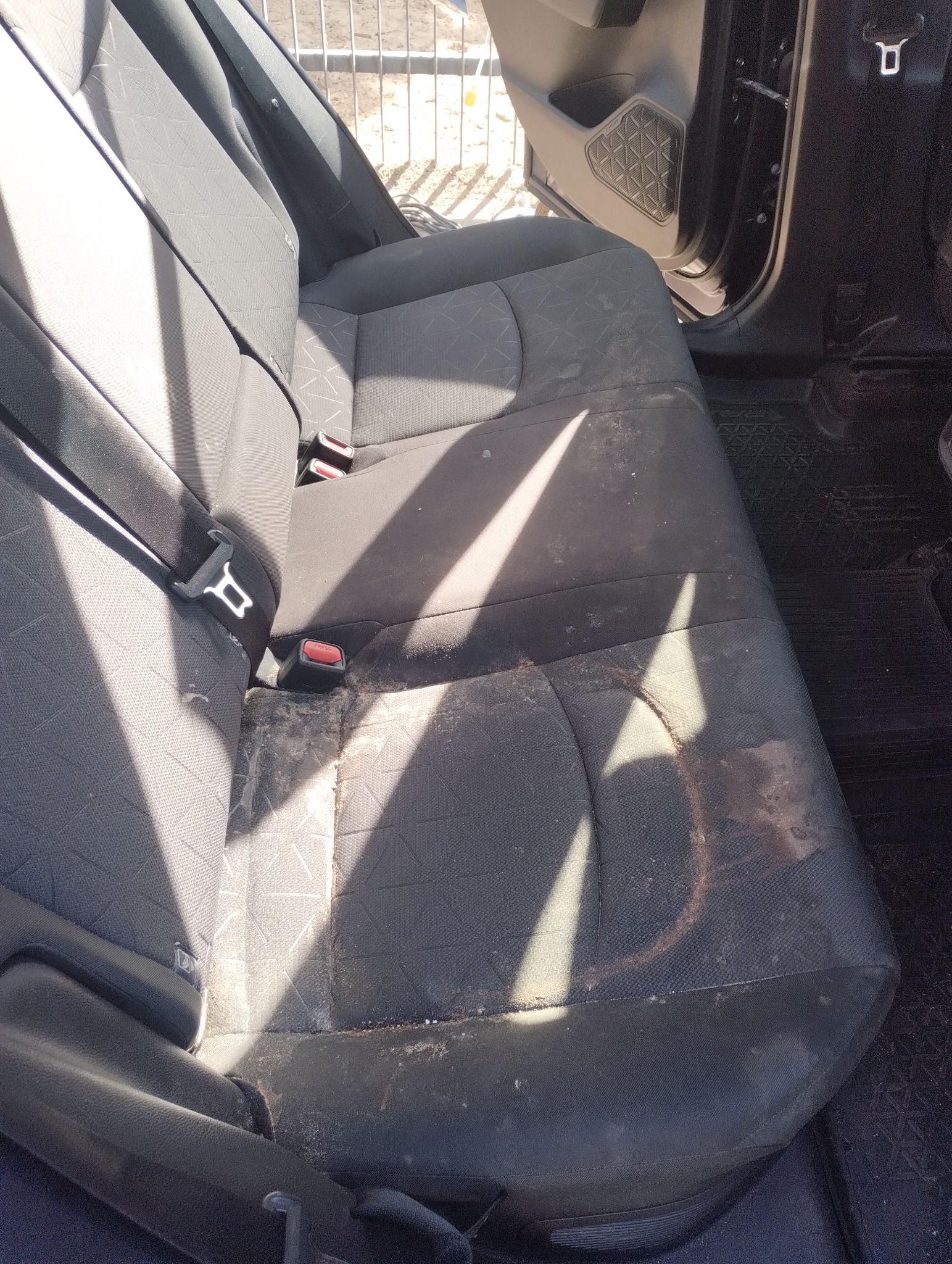 Bardzo zabrudzona, tylna kanapa samochodu z plamami i osadami. Widoczne pasy bezpieczeństwa i fragment drzwi. Potrzeba gruntownego czyszczenia tapicerki samochodowej.