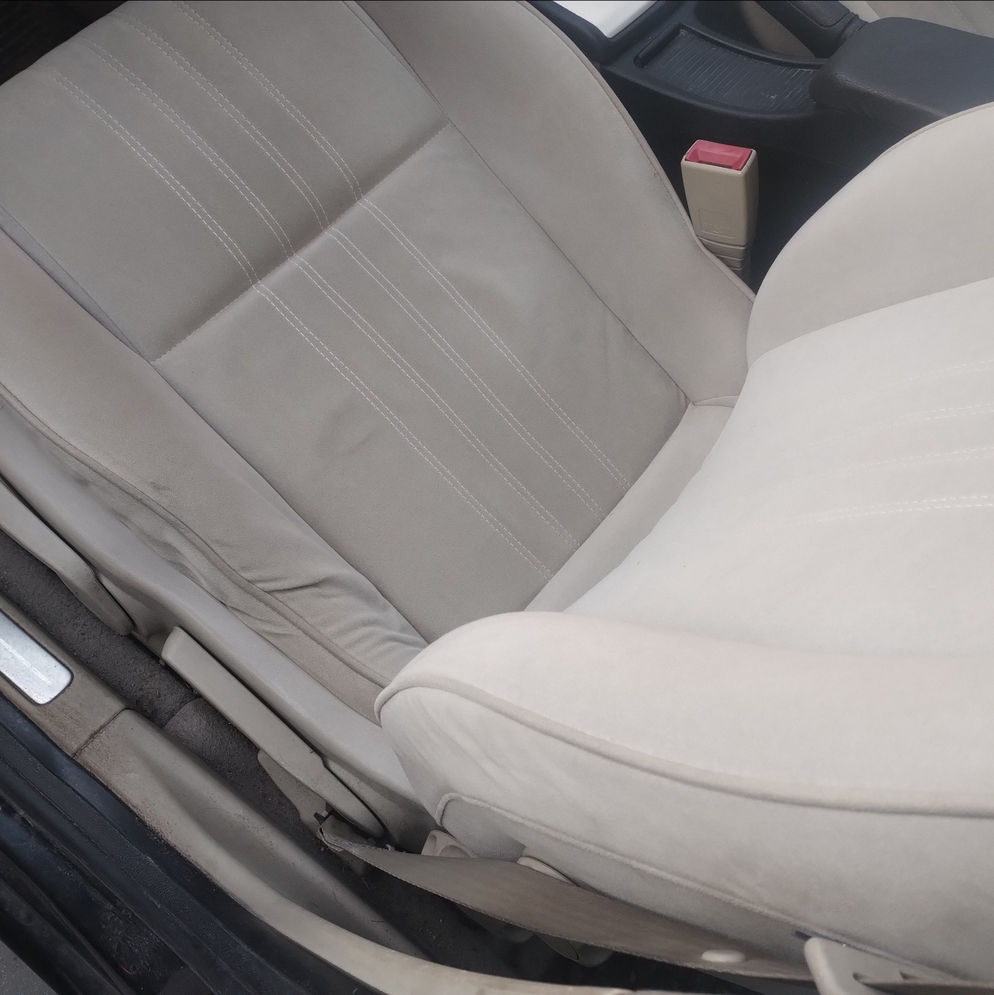 Jasne, tapicerowane siedzenie samochodowe z widocznymi przeszyciami i elementami wnętrza pojazdu. Widoczne zabrudzenia na tapicerce.