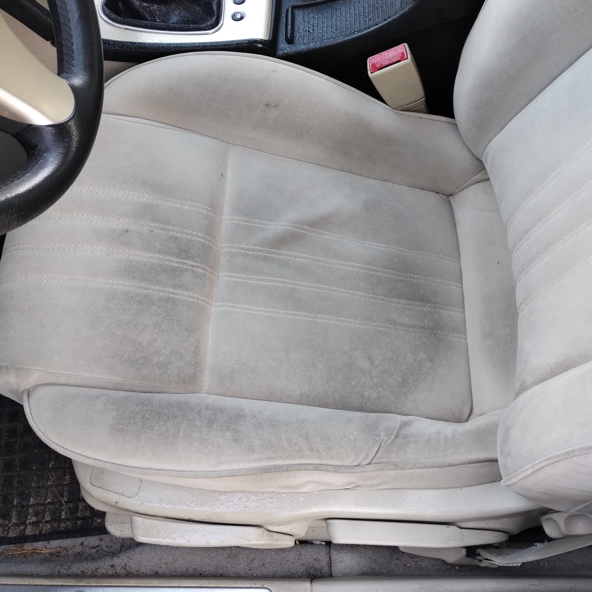 Brudny, jasny fotel samochodowy z plamami i zabrudzeniami na tapicerce. Widoczna część kierownicy i fragment deski rozdzielczej. Fotel wymaga czyszczenia.