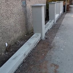 FREIS BUD - Surowy mur oporowy z betonowych elementów, świeżo po montażu, przylegający do ściany budynku z widoczną fakturą. Widok z poziomu chodnika.