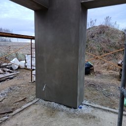 FREIS BUD - Surowy, szary filar z betonu w trakcie budowy domu jednorodzinnego, widoczny na tle krajobrazu z polami i wzgórzem. Element konstrukcyjny w stanie deweloperskim.