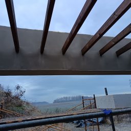 FREIS BUD - Szara elewacja z drewnianymi belkami, widok z dołu, rusztowanie w tle. Prace wykończeniowe na zewnątrz budynku w pochmurny dzień.