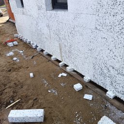 FREIS BUD - Ściana budynku w trakcie ocieplania, widoczne białe bloki styropianowe z szarymi plamkami, ułożone na drewnianej belce, z elementami instalacji elektrycznej w tle.