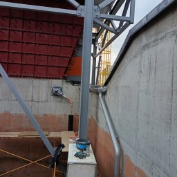 FREIS BUD - Konstrukcja stalowa przy betonowej ścianie z elementami elewacji w kolorze czerwonym. Widoczny fragment rynny i rusztowania. Prace budowlane w toku.