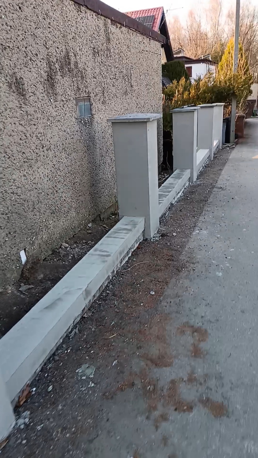 Surowy mur oporowy z betonowych elementów, świeżo po montażu, przylegający do ściany budynku z widoczną fakturą. Widok z poziomu chodnika.