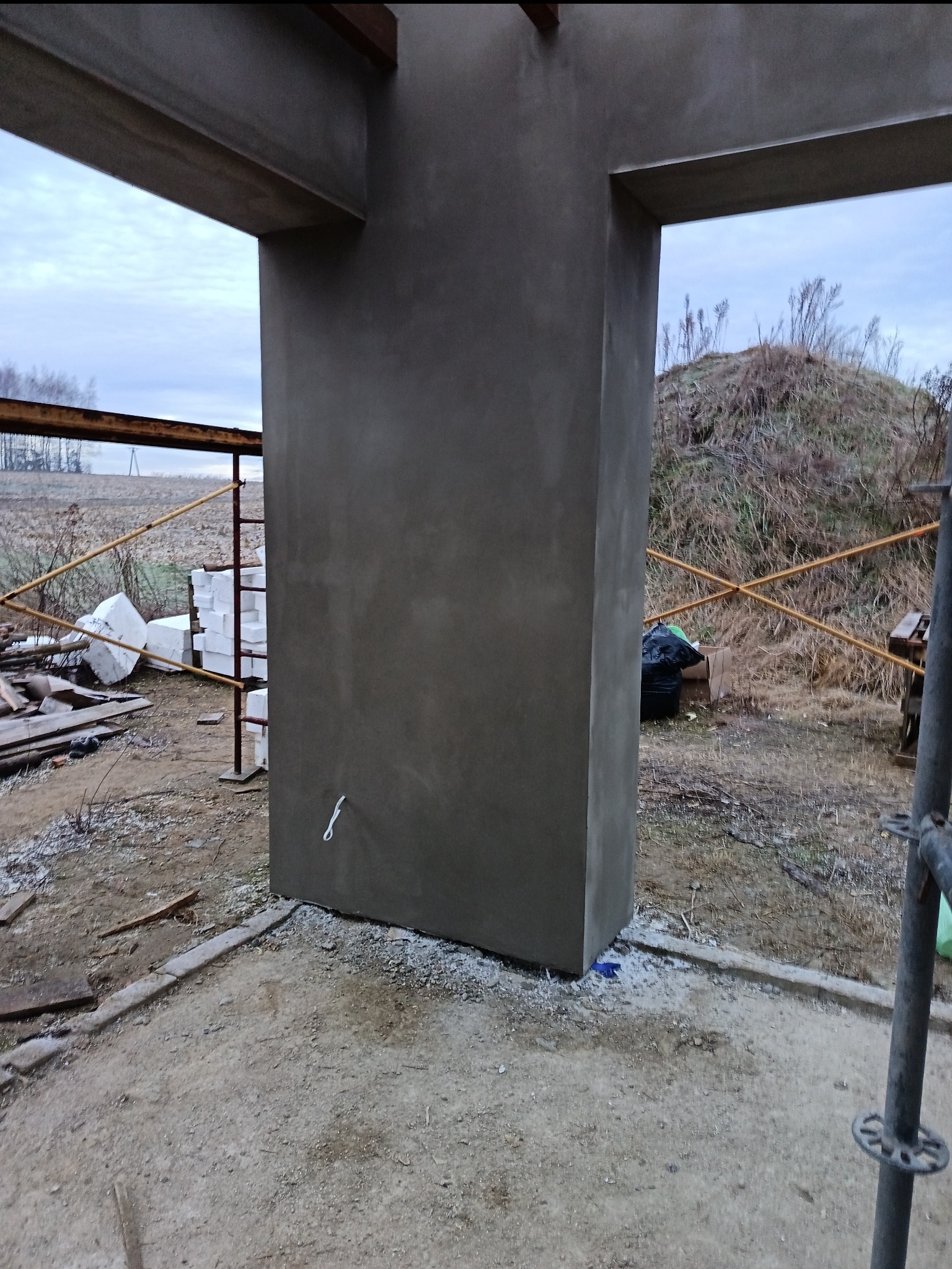 Surowy, szary filar z betonu w trakcie budowy domu jednorodzinnego, widoczny na tle krajobrazu z polami i wzgórzem. Element konstrukcyjny w stanie deweloperskim.