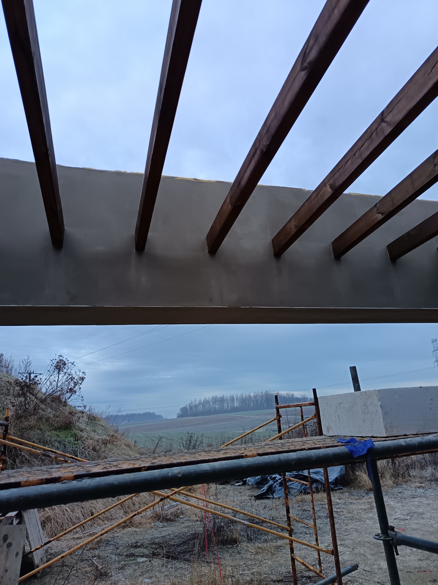 Szara elewacja z drewnianymi belkami, widok z dołu, rusztowanie w tle. Prace wykończeniowe na zewnątrz budynku w pochmurny dzień.