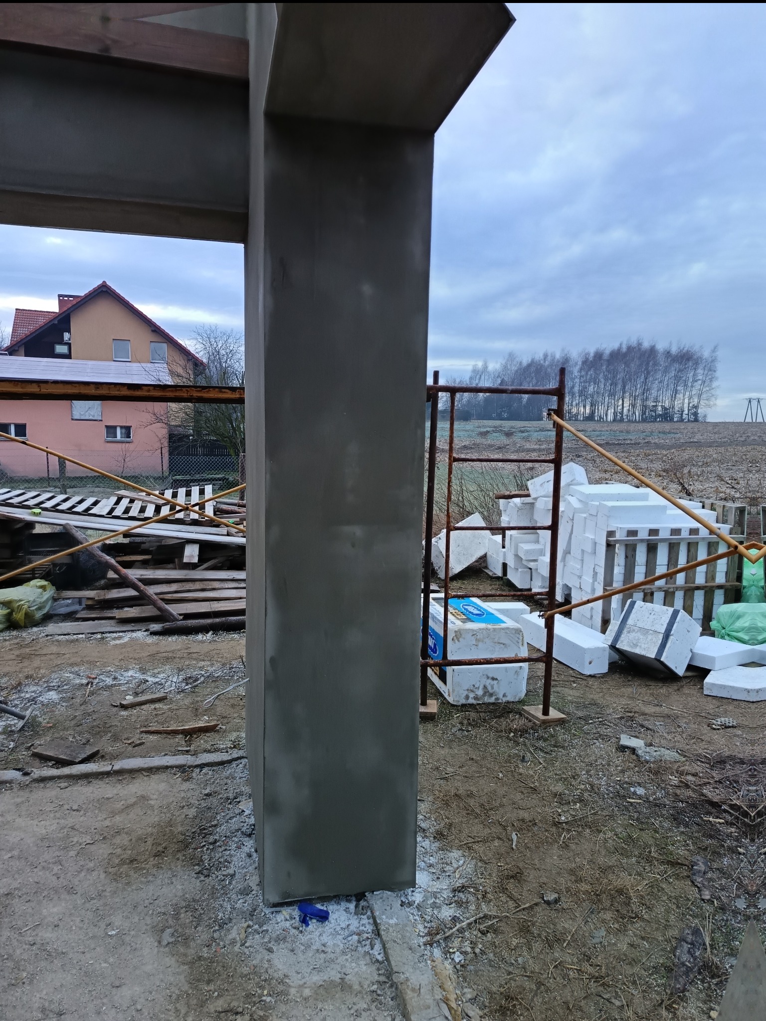 Wznoszenie konstrukcji budynku: otynkowany filar na tle placu budowy z materiałami izolacyjnymi i rusztowaniem. Widok na dom i drzewa w tle.