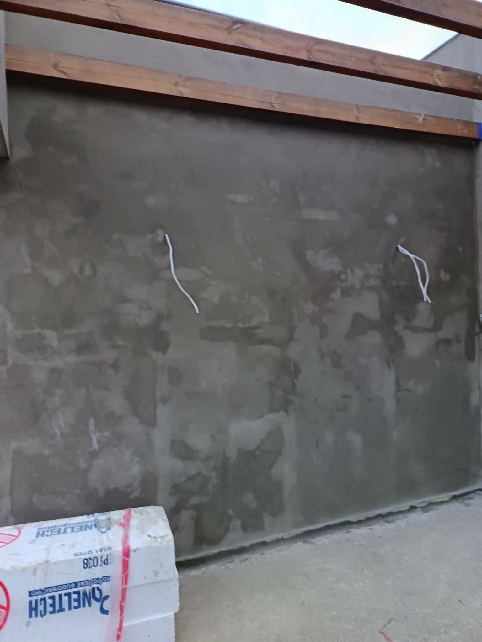 Szara, surowa ściana z wystającymi przewodami elektrycznymi, częściowo zakryta przez białe płyty styropianowe. Widoczne drewniane elementy konstrukcyjne pod sufitem.