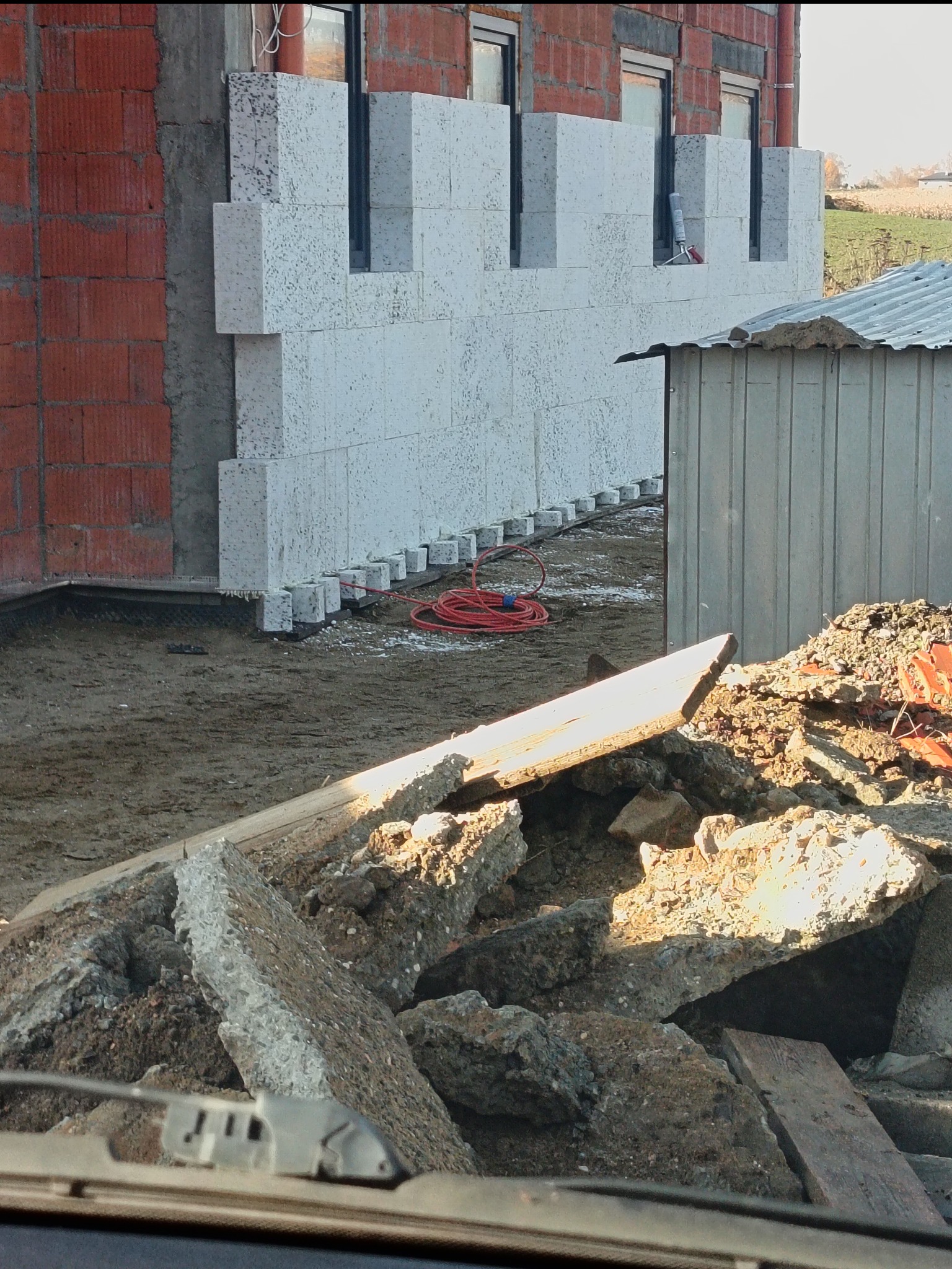 Budowa domu: fragment ściany z białych bloczków, widoczne okna i czerwona cegła. Na pierwszym planie gruz i ziemia. W tle pole i budynki.