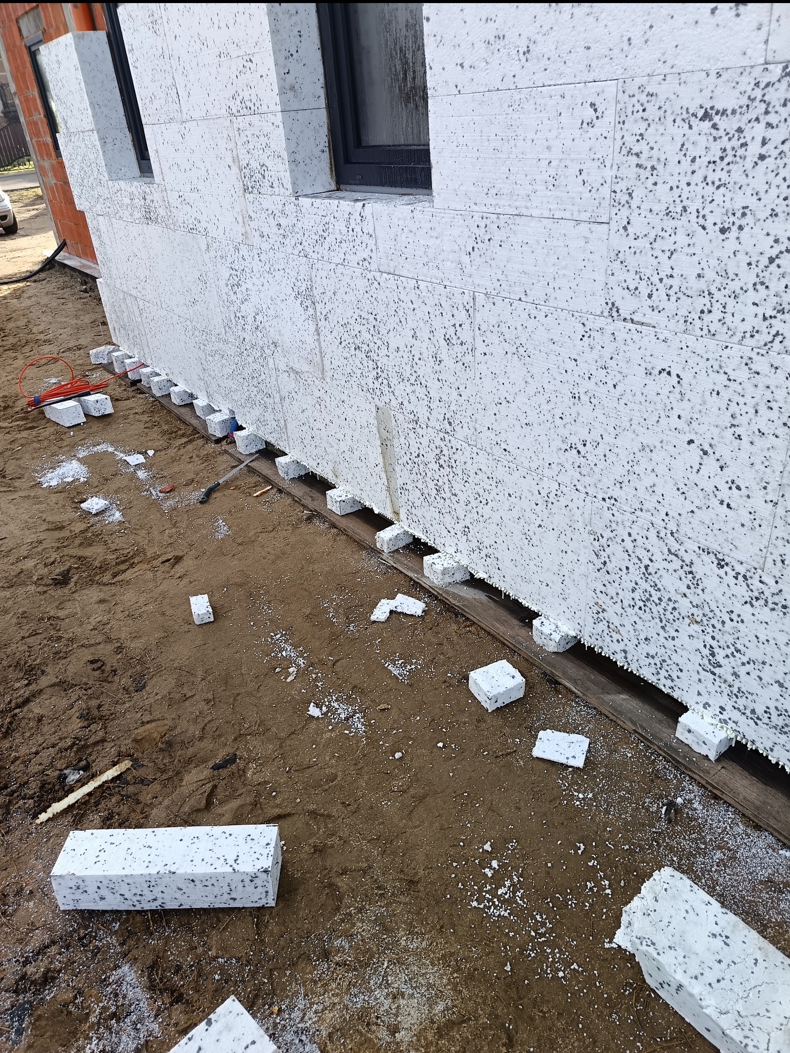 Ściana budynku w trakcie ocieplania, widoczne białe bloki styropianowe z szarymi plamkami, ułożone na drewnianej belce, z elementami instalacji elektrycznej w tle.