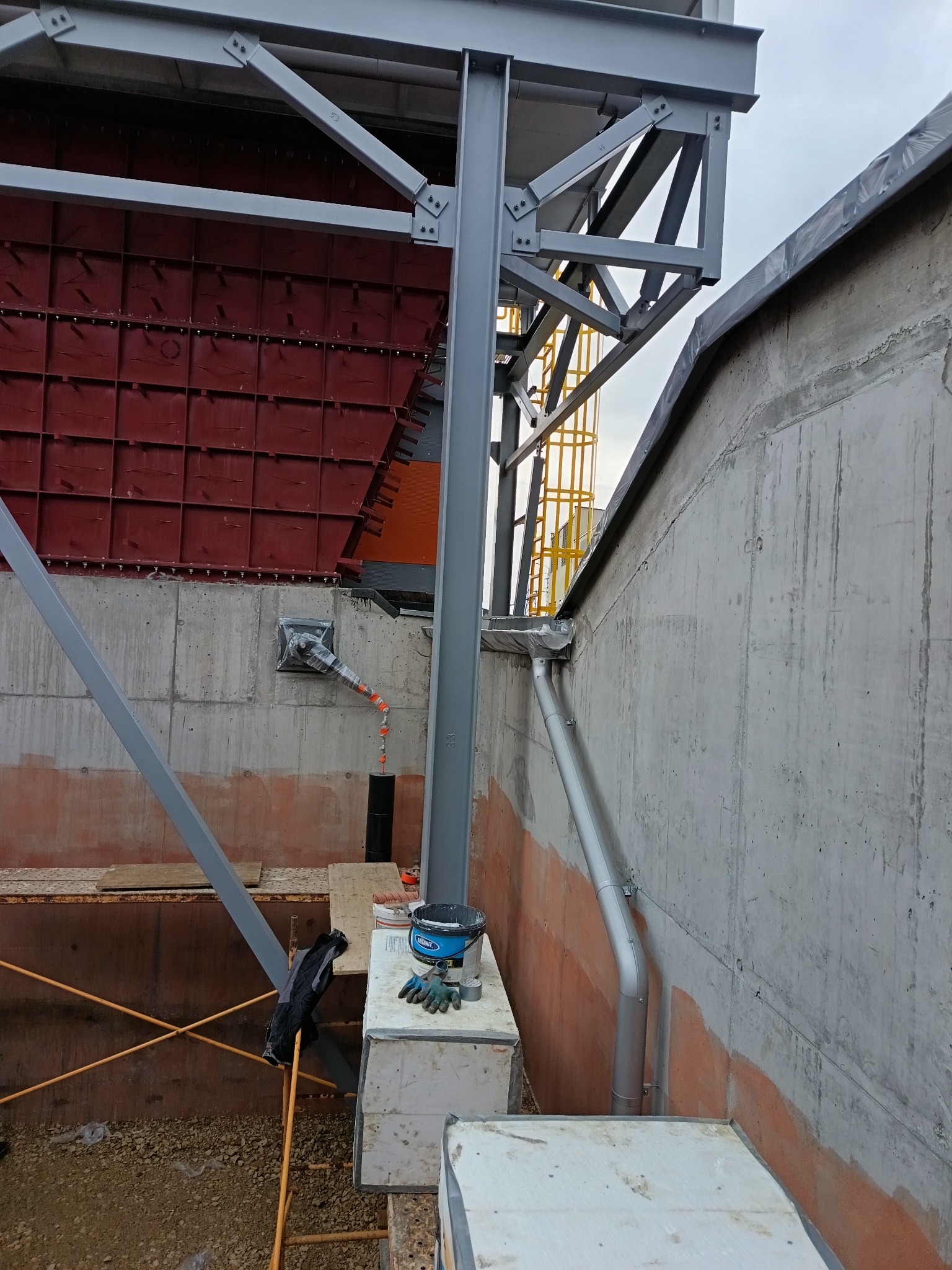 Konstrukcja stalowa przy betonowej ścianie z elementami elewacji w kolorze czerwonym. Widoczny fragment rynny i rusztowania. Prace budowlane w toku.
