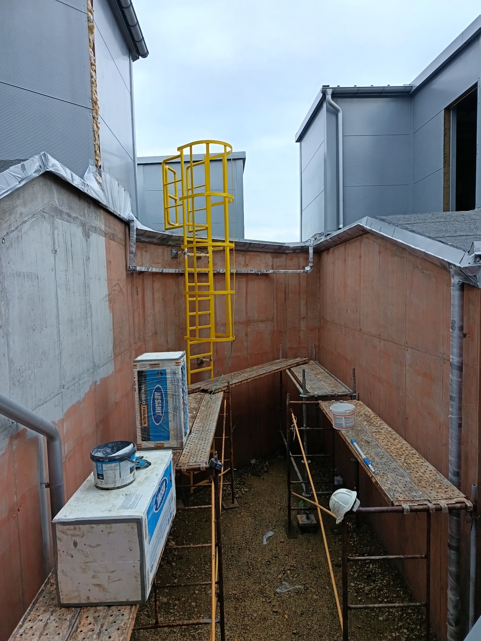 Przygotowania do prac na elewacji budynku: rusztowania, drabina, materiały izolacyjne i puszka z farbą. Widok z góry na plac budowy.