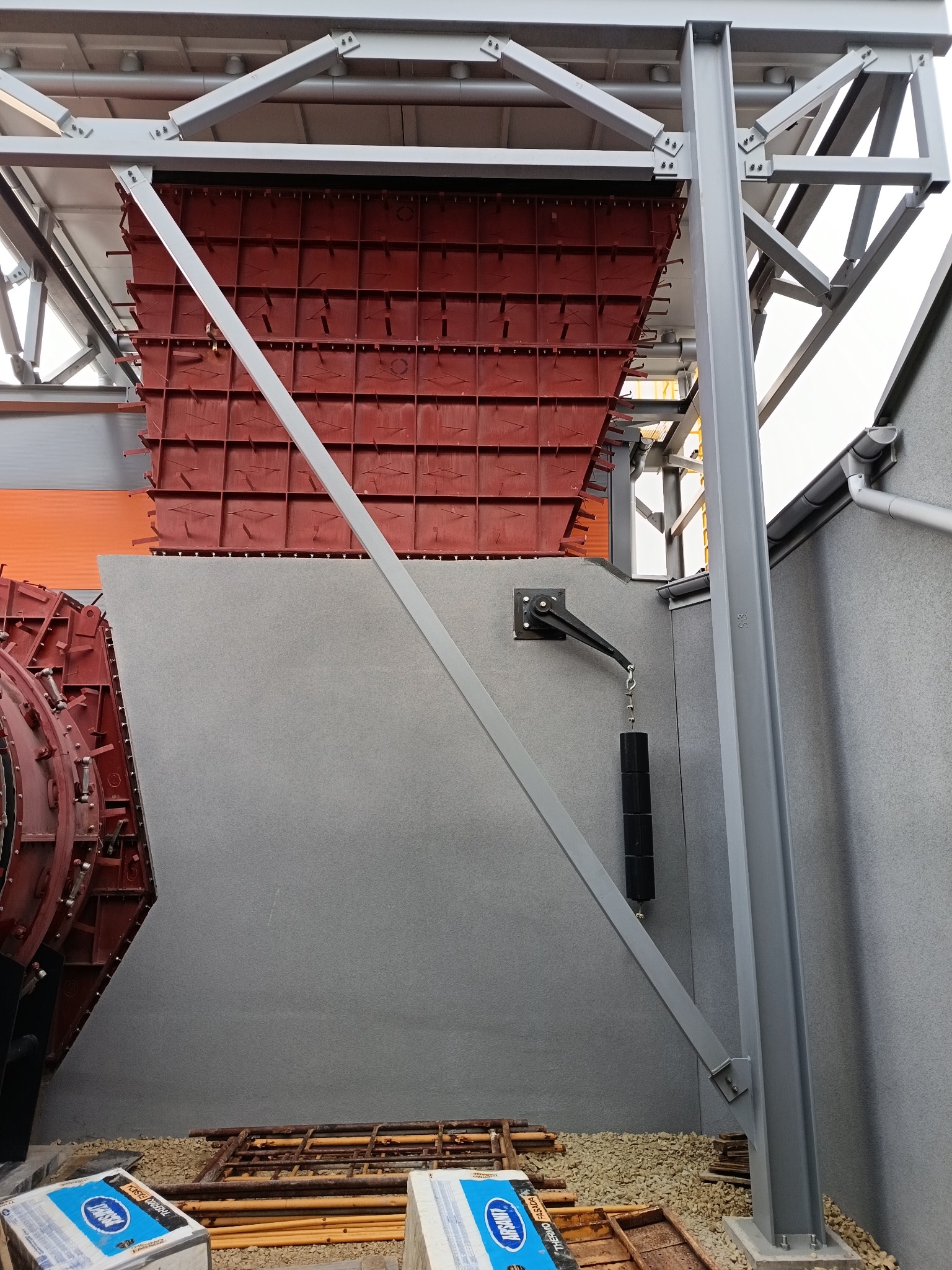 Szara ściana z elementami konstrukcyjnymi i czerwonymi formami szalunkowymi. Widoczne materiały budowlane i rusztowania. Detal z budowy, surowy stan.