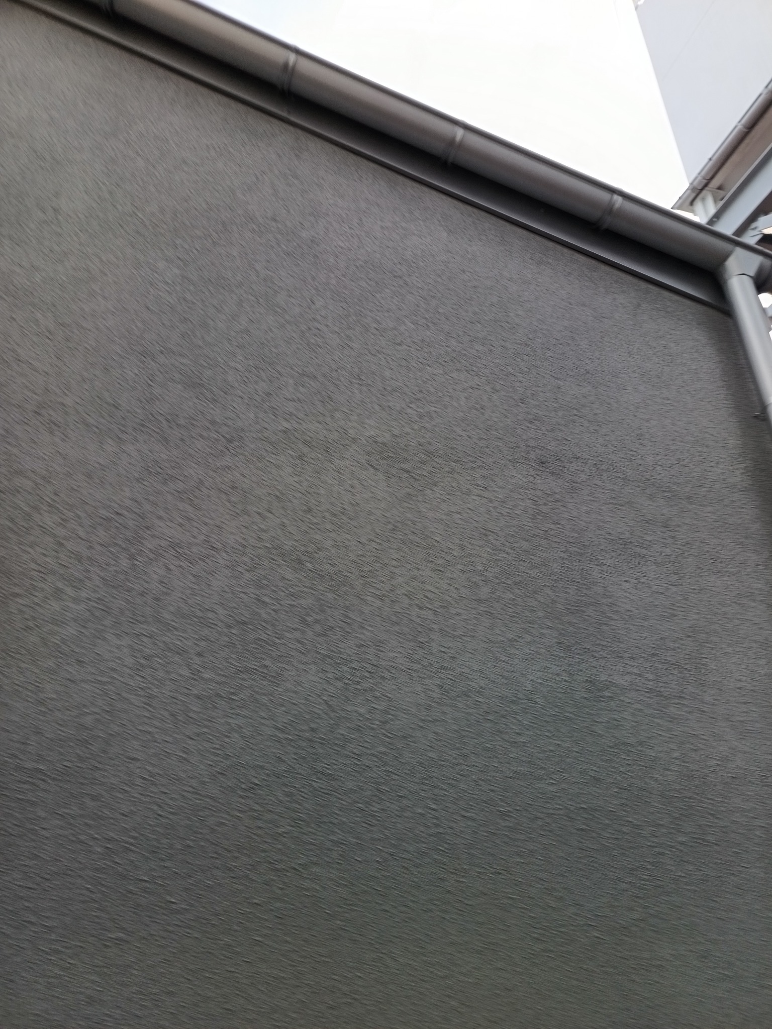 Szara, teksturowana elewacja budynku z widoczną rynną. Ujęcie z dołu, podkreślające fakturę ściany i detale orynnowania.