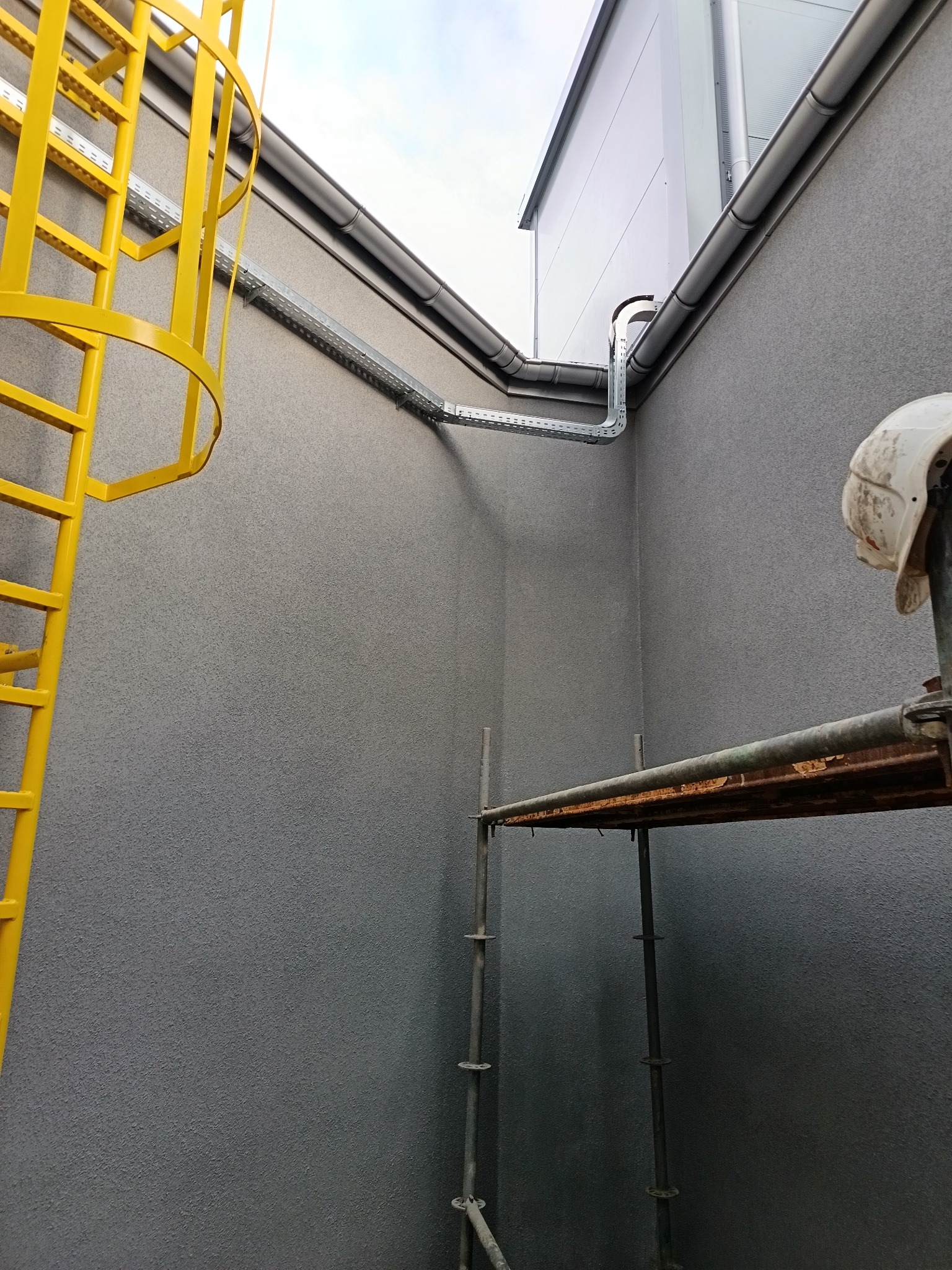 Szara elewacja budynku z widoczną żółtą drabiną, rynnami i korytami kablowymi. Fragment rusztowania z kaskiem budowlanym w prawym górnym rogu.