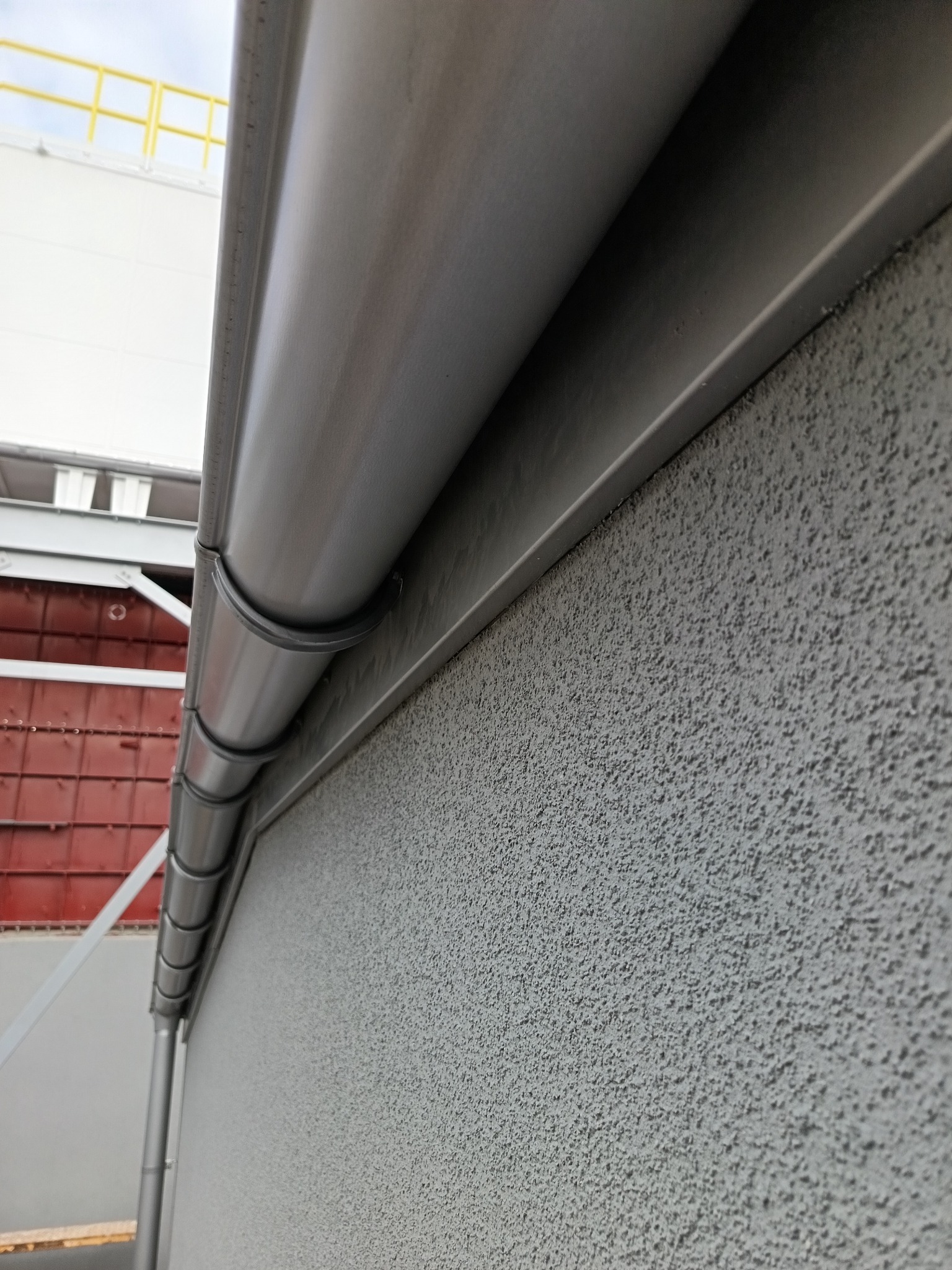 Szary budynek z zamontowaną rynną. Widoczna faktura elewacji i elementy konstrukcyjne budynku w tle. Ujęcie z dołu, podkreślające detale wykończenia.