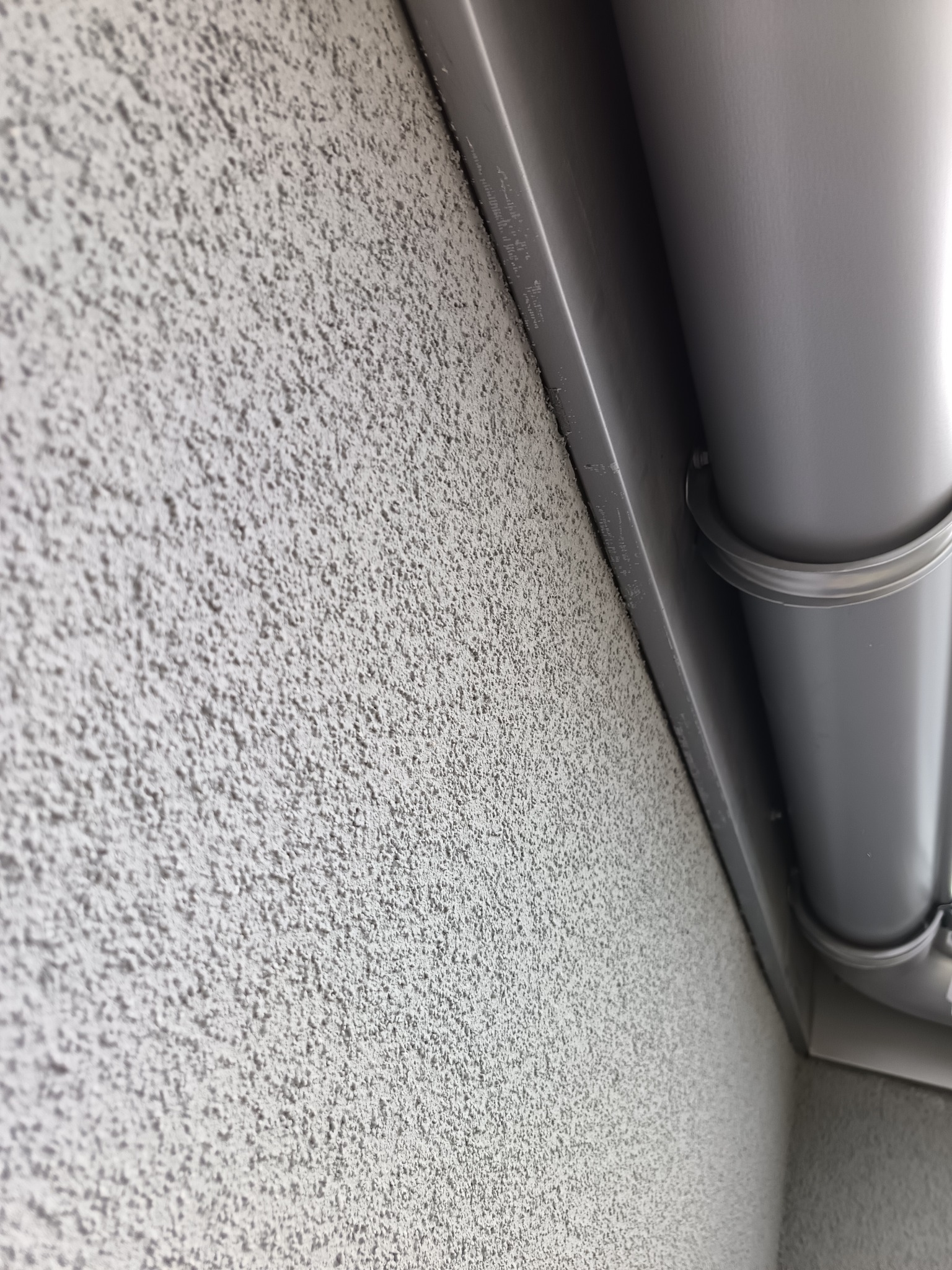 Szara elewacja z fakturą baranka i fragmentem orynnowania. Detal wykończenia ściany zewnętrznej budynku z bliska, widoczny system odprowadzania wody.