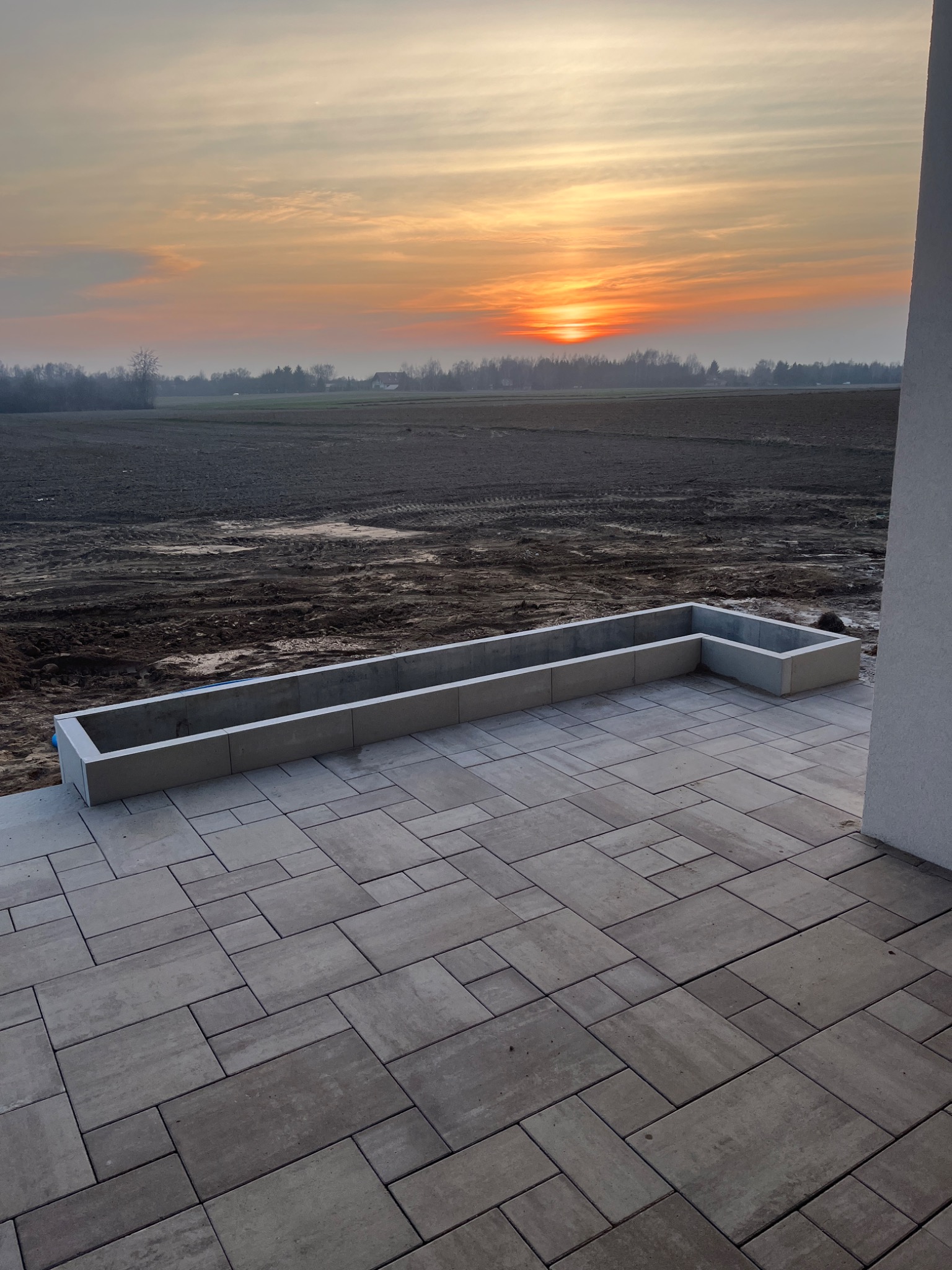 Ułożona kostka brukowa w odcieniach szarości, z betonową donicą w tle i zachodem słońca. Widok z perspektywy narożnika budynku.
