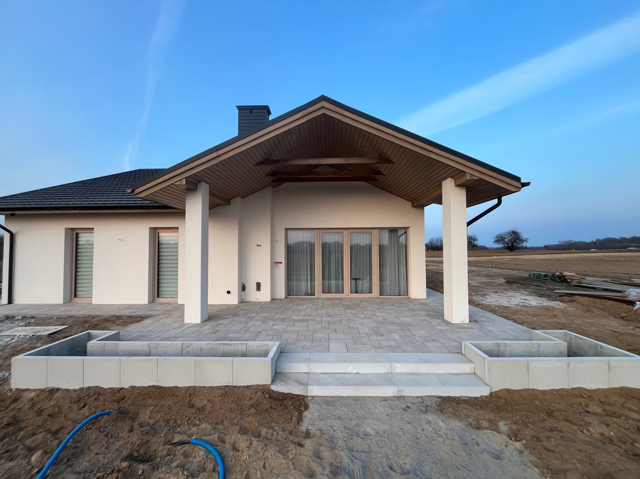 Nowoczesny dom z tarasem wyłożonym jasną kostką brukową. Widoczne schody i rabaty kwiatowe w surowym stanie. Tło stanowi błękitne niebo i pole.