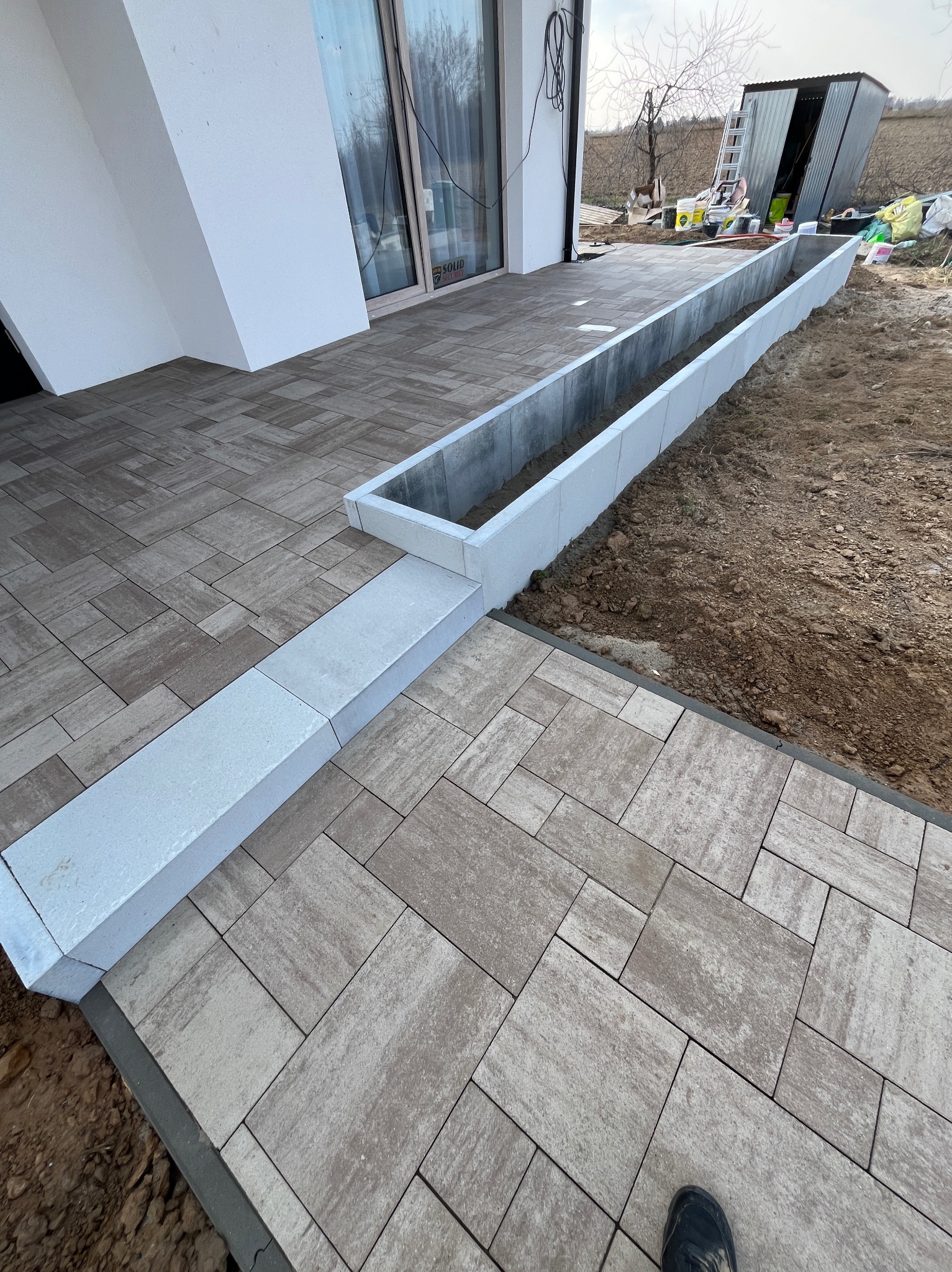 Ułożona kostka brukowa w odcieniach beżu i brązu, tworząca ścieżkę z obrzeżami. Widoczny fragment budynku i ziemia. Narożny kadr.