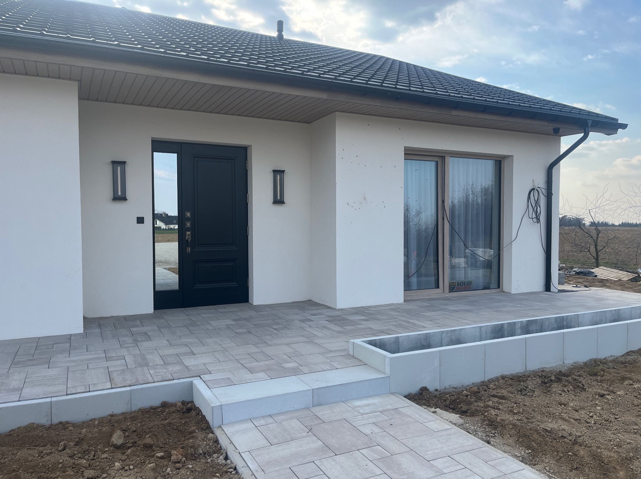 Nowoczesny podjazd z jasnoszarej kostki brukowej prowadzący do domu z czarnymi drzwiami i oknami. Widoczne schody i obrzeża z kostki. Minimalistyczny design.