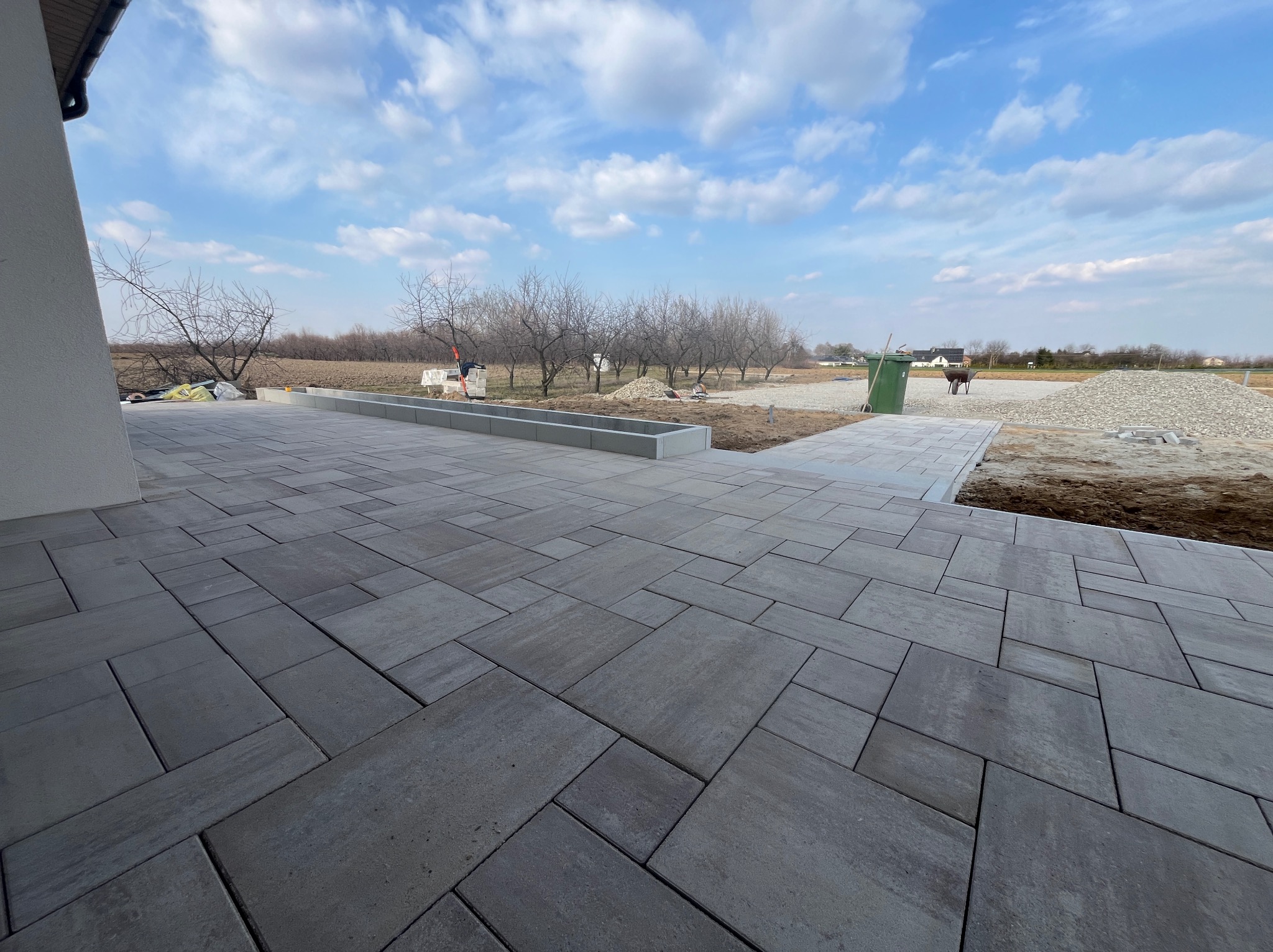 Ułożona, szara kostka brukowa na posesji domu jednorodzinnego. Widoczne przygotowane podłoże i elementy krajobrazu. Słoneczny dzień, błękitne niebo z chmurami.