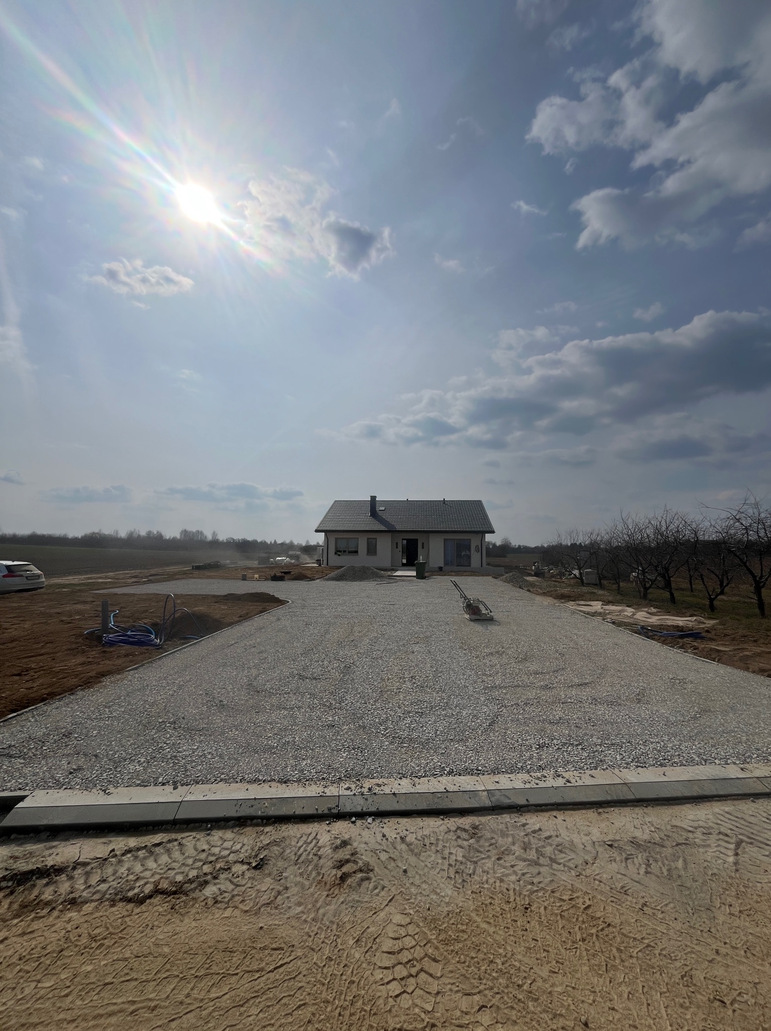 Szeroki kadr na podjazd z szarego kruszywa prowadzący do nowoczesnego domu jednorodzinnego. W tle błękitne niebo z chmurami i intensywne słońce.