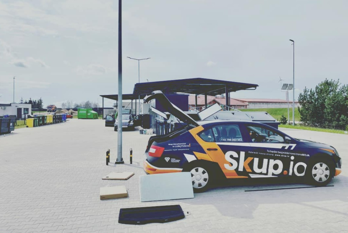 Auto z logo skup.io na parkingu, otwarty bagażnik, elementy wyposażenia obok. Usługa skupu nieruchomości w całej Polsce, szybka transakcja.