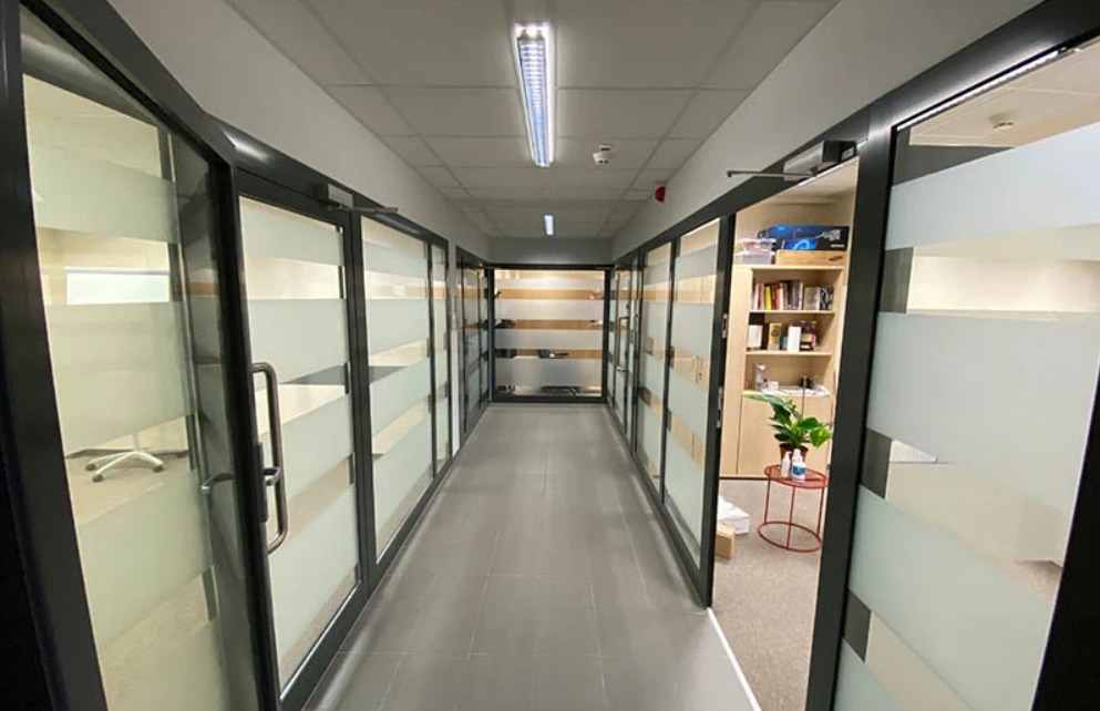 Korytarz biurowy z przeszklonymi biurami po bokach, z matowymi pasami na szybach. Widoczne biurka, krzesła i regały. Szara podłoga, sufit z oświetleniem liniowym.