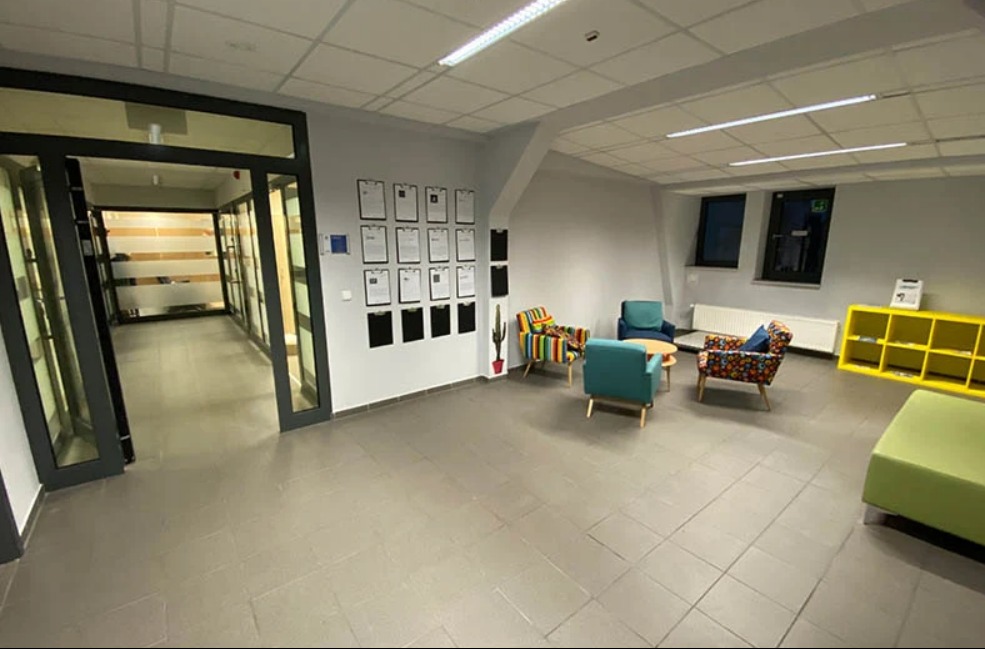 Jasne wnętrze biura nieruchomości z kolorowymi fotelami i dokumentami na ścianie. Widoczne okna i przeszklone drzwi wejściowe.