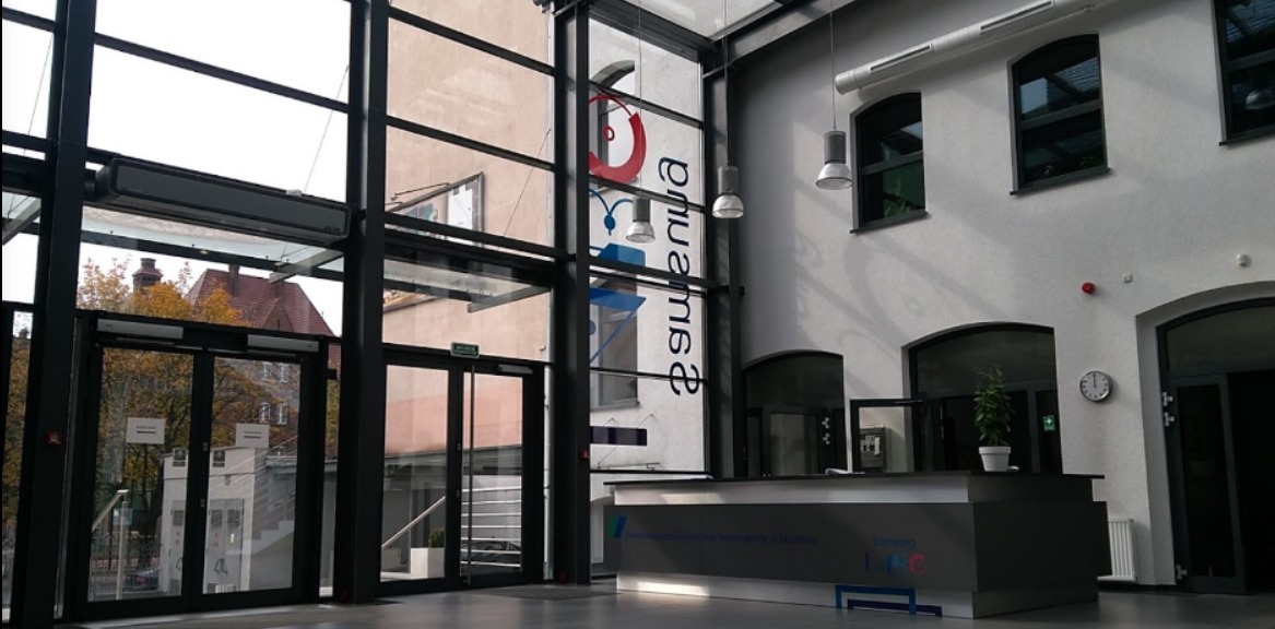 Nowoczesne wnętrze biura z recepcją, przeszklonym wejściem i logo firmy na ścianie. Widoczne elementy architektoniczne i oświetlenie.