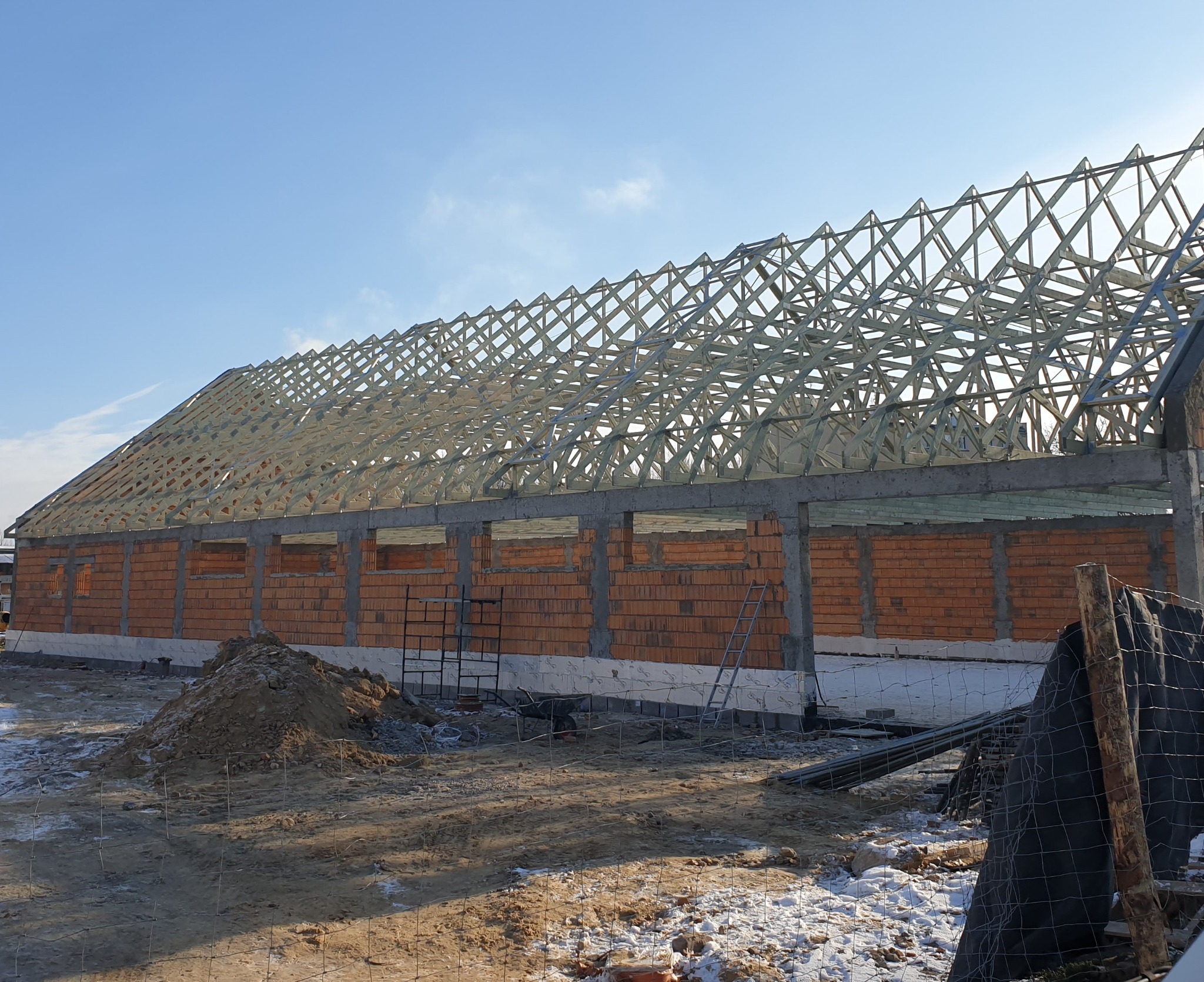 Budowa hali: surowa konstrukcja dachu z drewna na murowanych ścianach z pustaków. Widoczne rusztowania i ziemia. Słoneczny dzień.