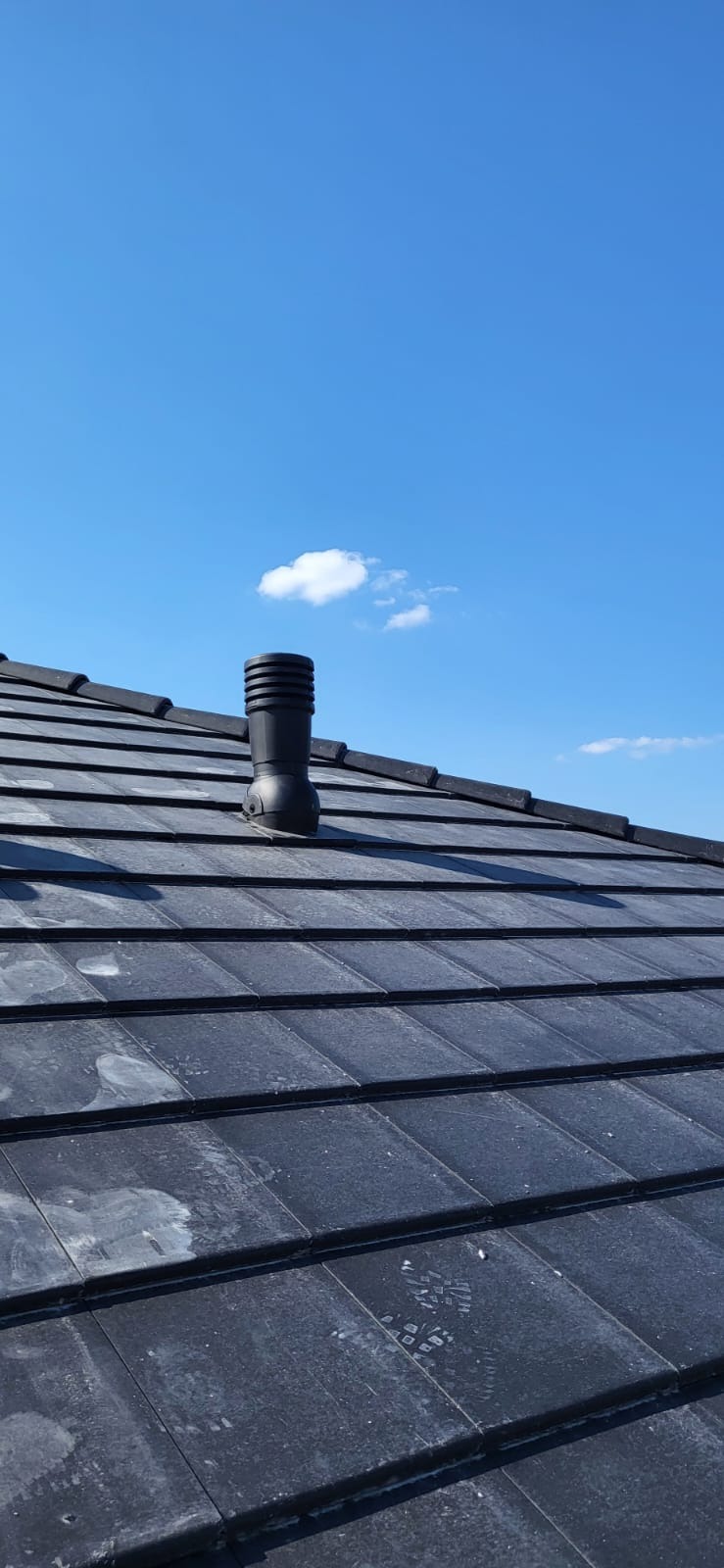 Czarny kominek wentylacyjny na dachu z ciemnej dachówki ceramicznej, na tle błękitnego nieba z chmurami. Widoczne ślady na dachówkach.