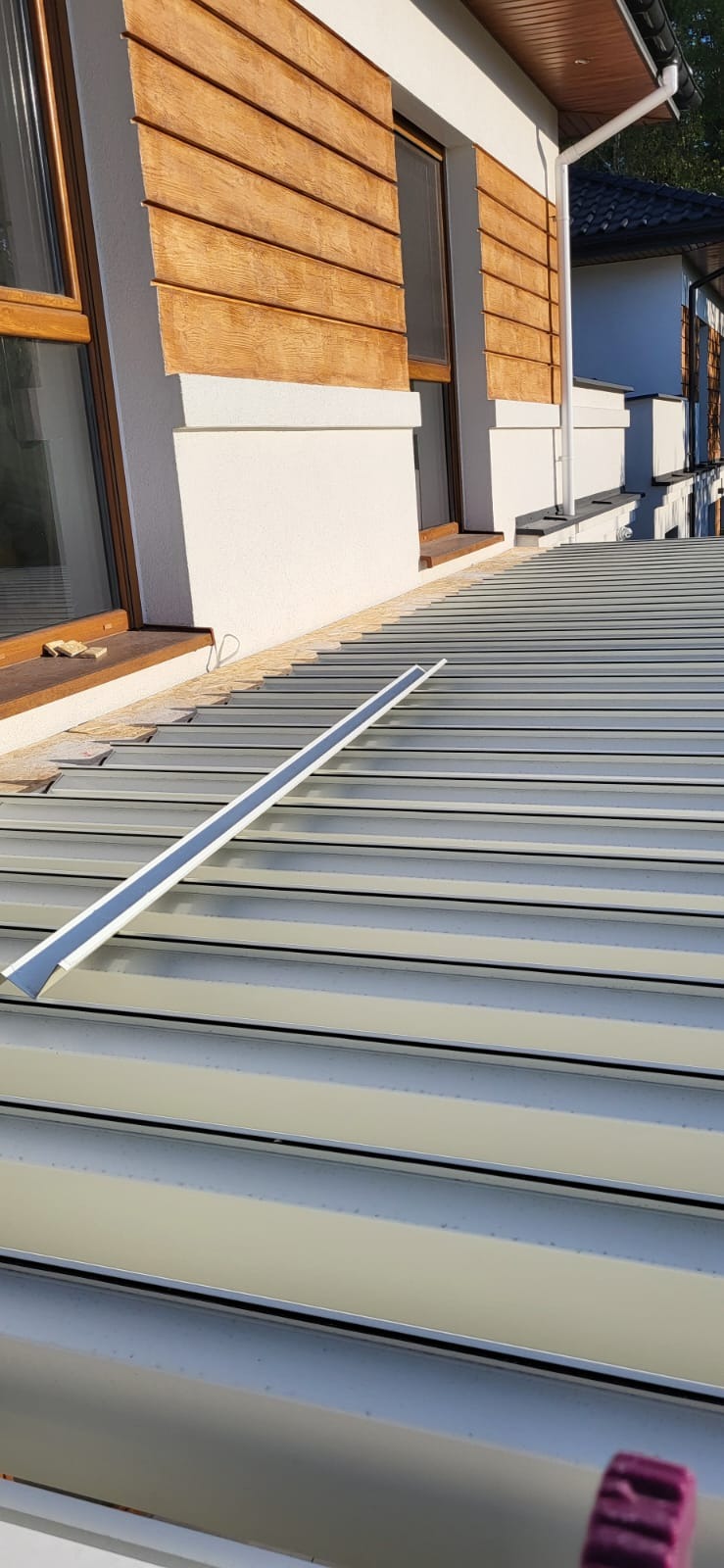 Jasnoszary dach z blachy trapezowej w trakcie montażu, widoczne elementy mocujące. Budynek z drewnianą elewacją i oknami w tle. Ujęcie z góry, w słoneczny dzień.