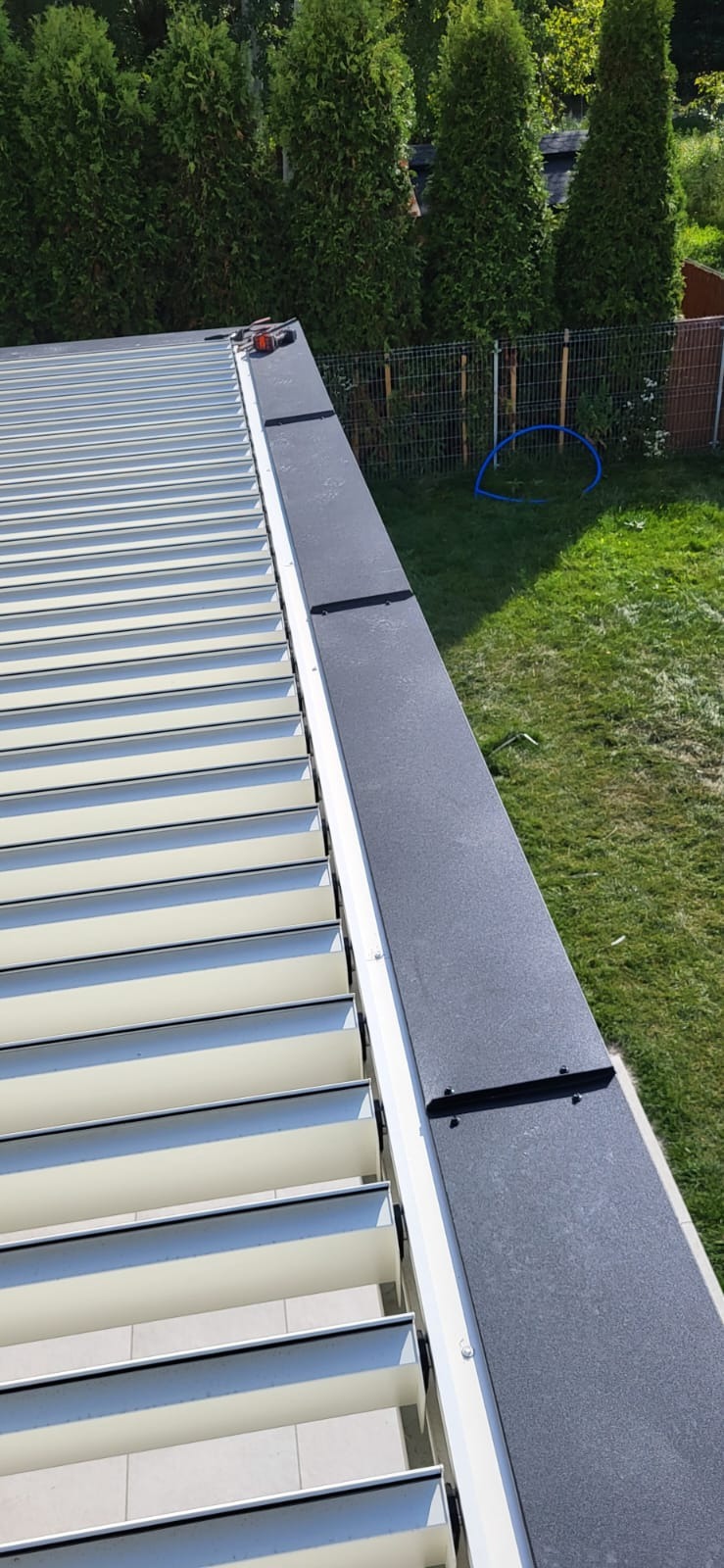 Nowoczesny dach z jasnymi panelami i ciemnymi obróbkami blacharskimi, widok z góry na tle zielonego trawnika i drzew. Detal wykończenia dachu.