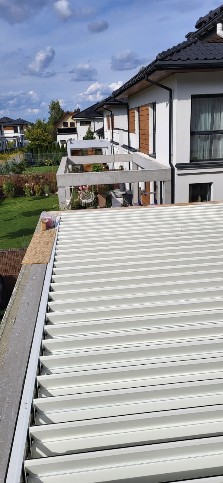 Białe, poziome panele dachowe z bliska, z widocznym fragmentem domu z brązowymi akcentami i zielonym ogrodem w tle, w słoneczny dzień.