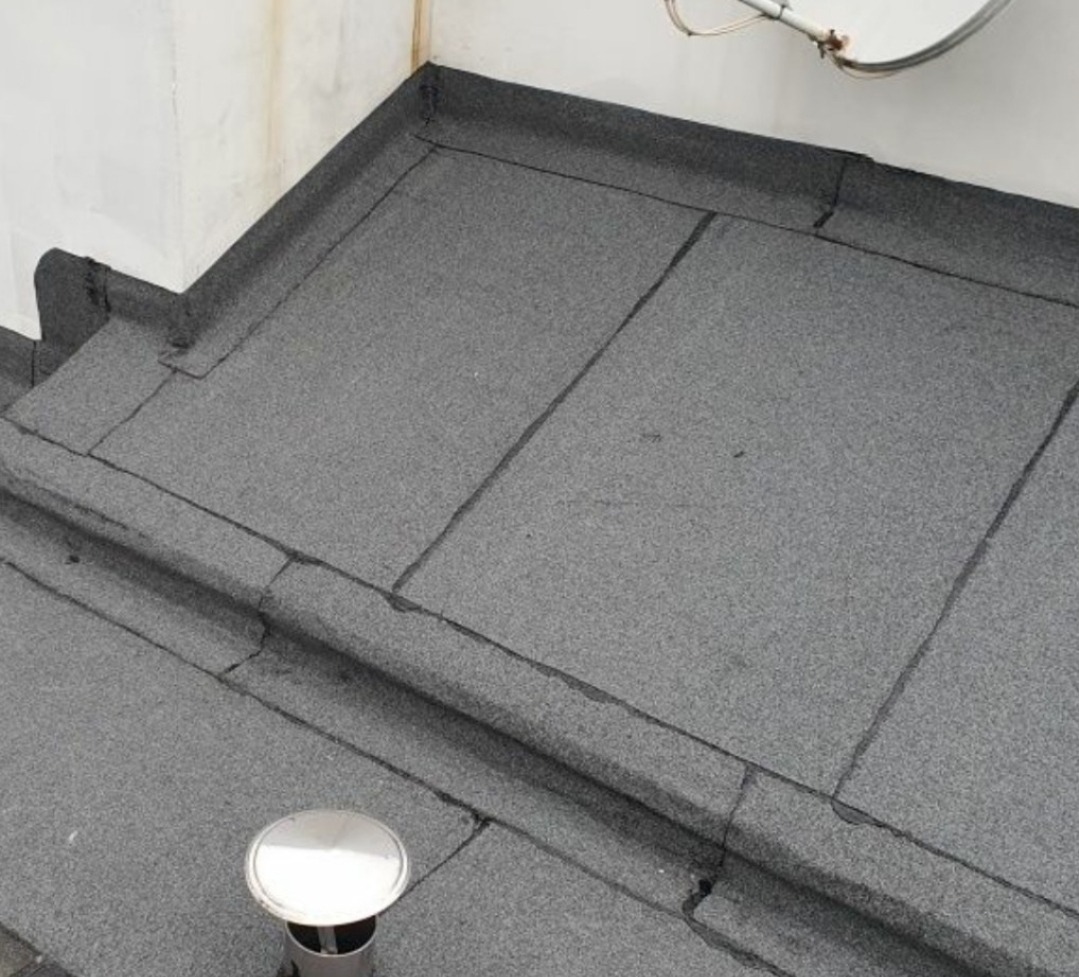 Szczelny, płaski dach pokryty papą termozgrzewalną z kominkiem wentylacyjnym i anteną satelitarną w rogu. Widok z góry.