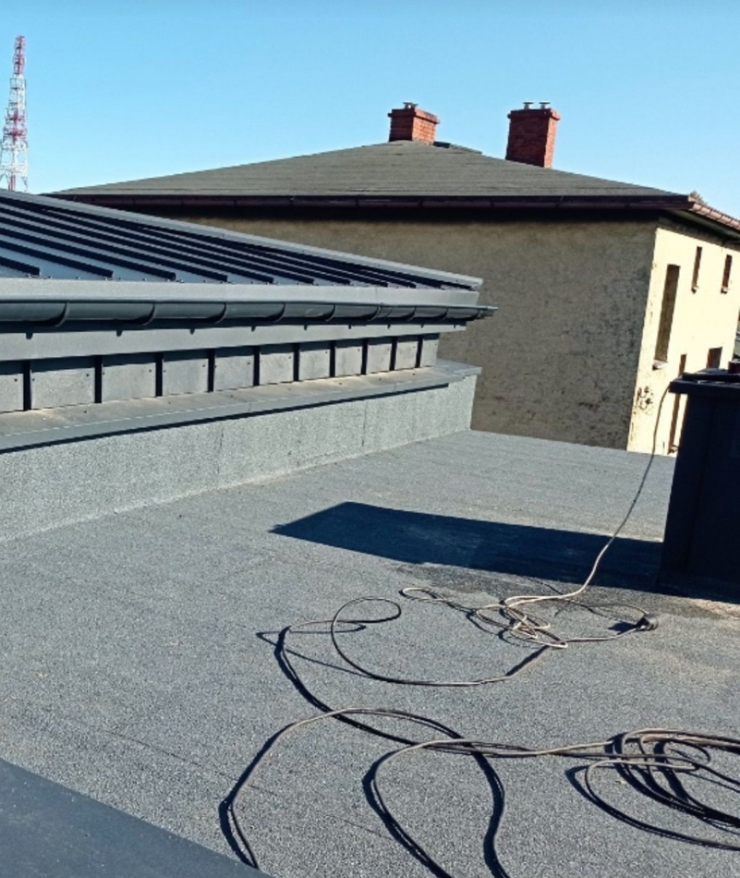 Szary dach pokryty papą z nową, ciemną rynną i obróbką blacharską. W tle budynek z ceglanymi kominami i wieża telekomunikacyjna na tle błękitnego nieba.