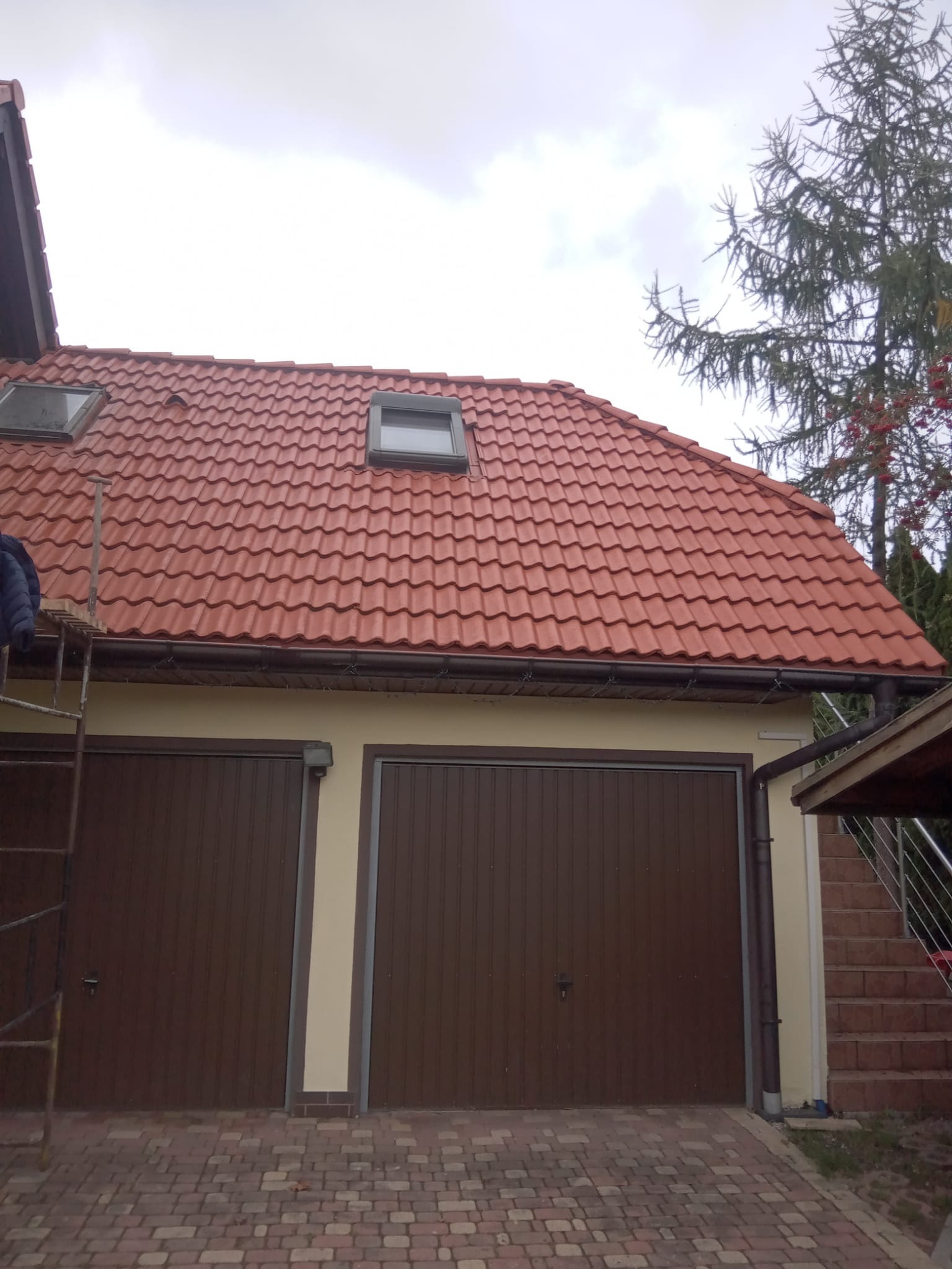 Czerwony dach z dachówką ceramiczną i oknami połaciowymi nad garażem z dwiema bramami. Widoczna drabina i fragment drzewa. Pochmurne niebo w tle.