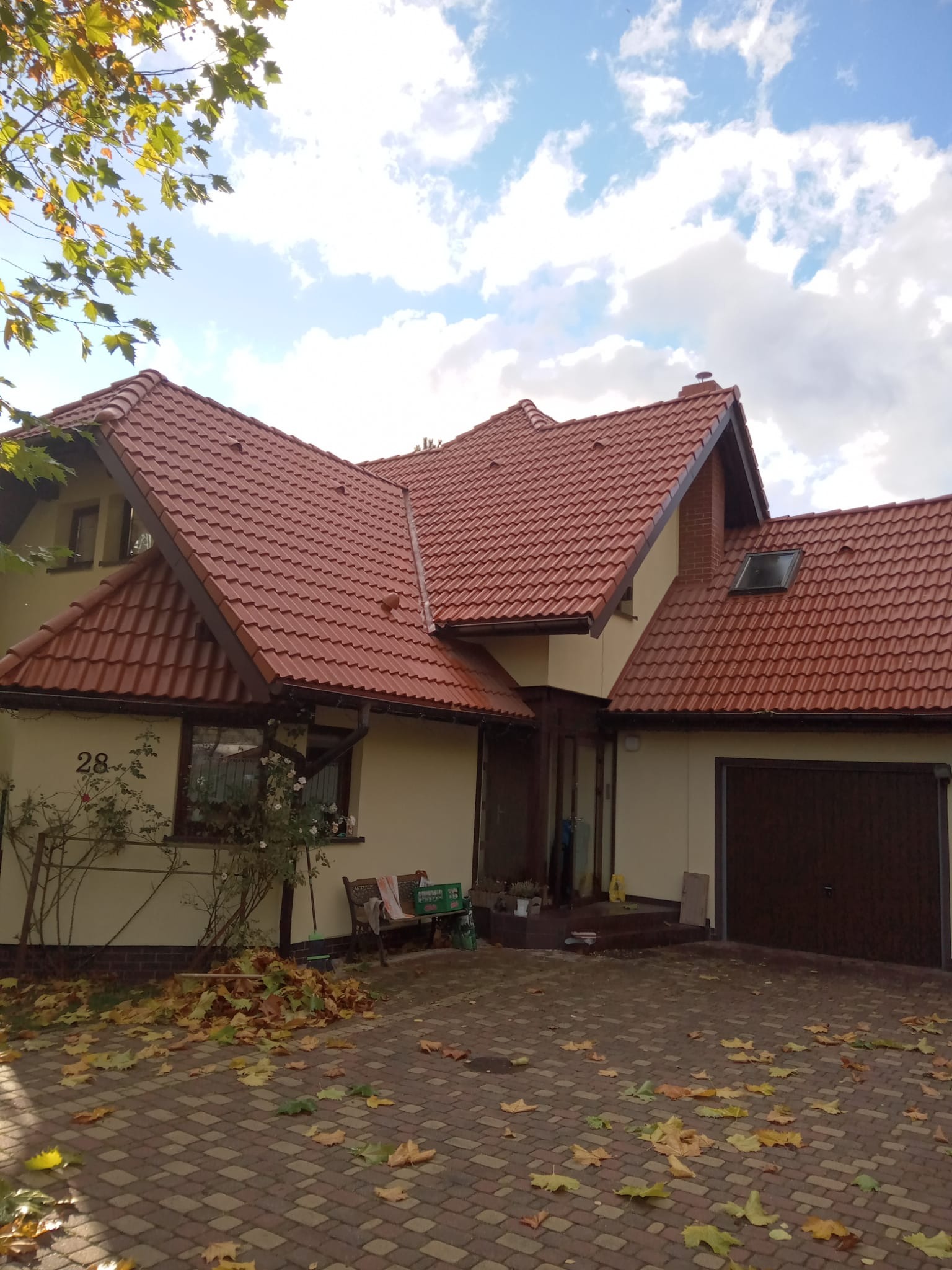 Dom jednorodzinny z nowym, czerwonym dachem z dachówki ceramicznej. Widoczne detale wykończenia, rynny i obróbki blacharskie. Jesienne liście na brukowanym podjeździe.