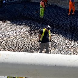 SP WARSZAWA SPÓŁKA Z OGRANICZONĄ ODPOWIEDZIALNOŚCIĄ - Pracownik w kamizelce odblaskowej układa zbrojenie na budowie. Widoczna siatka z prętów zbrojeniowych i fragmenty innych osób w tle. Praca wre.